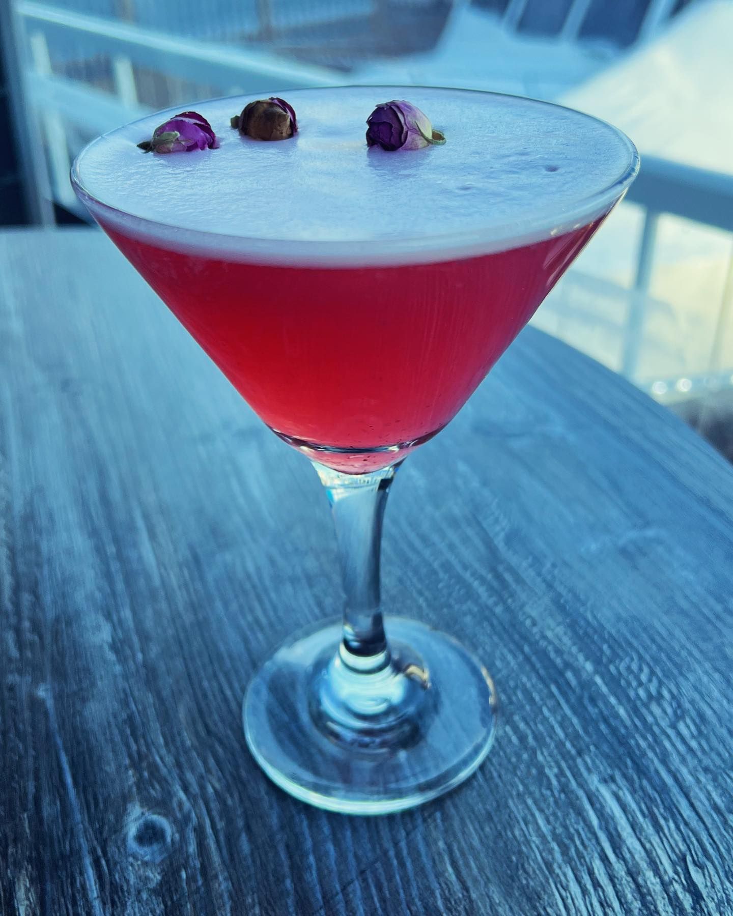 Pink cocktail in a martini glass, garnished with flowers, on a wooden surface.