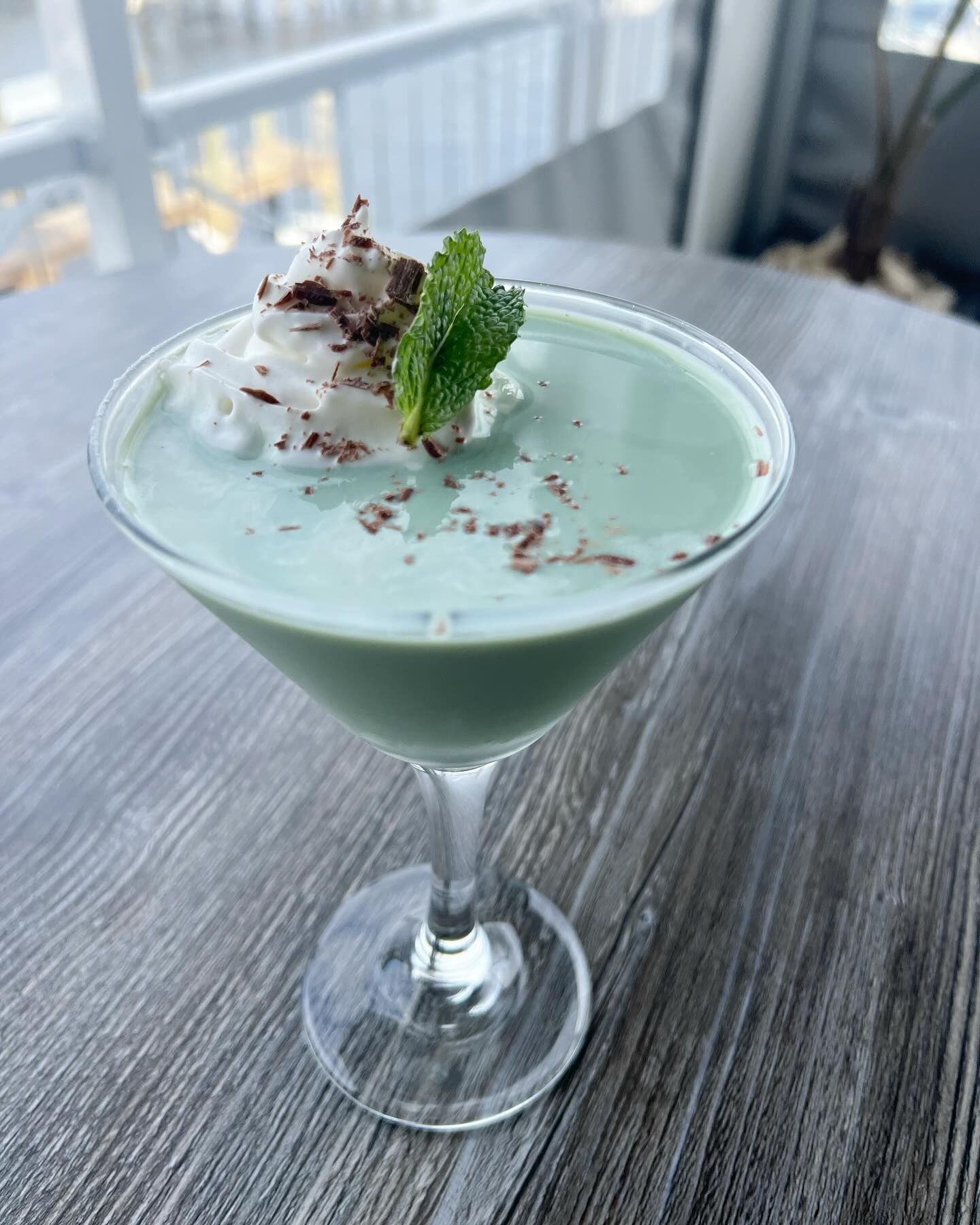 Green cocktail in a martini glass, topped with whipped cream, mint, and chocolate shavings on a gray table.