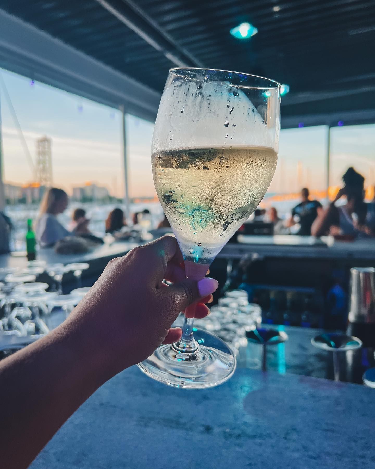 Hand holding a champagne glass at a waterfront bar, bubbles visible, people in background, sunset.