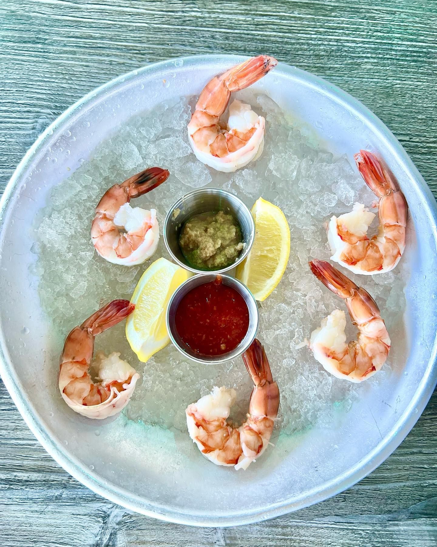 Shrimp cocktail on ice with lemon wedges and two dipping sauces in silver cups on a plate.