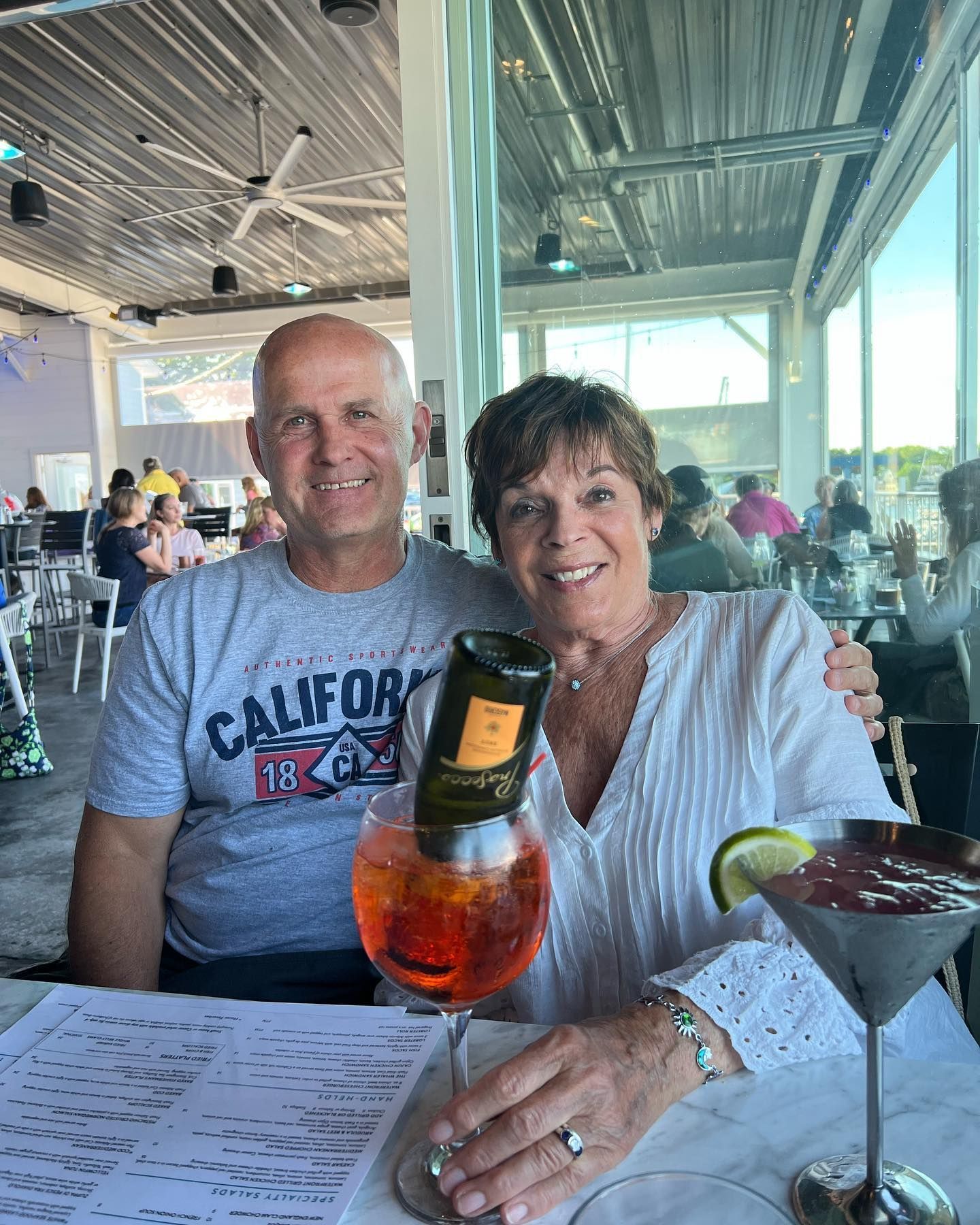 Couple seated at a restaurant table, smiling. Aperol spritz with a wine bottle and a cocktail in front of them.
