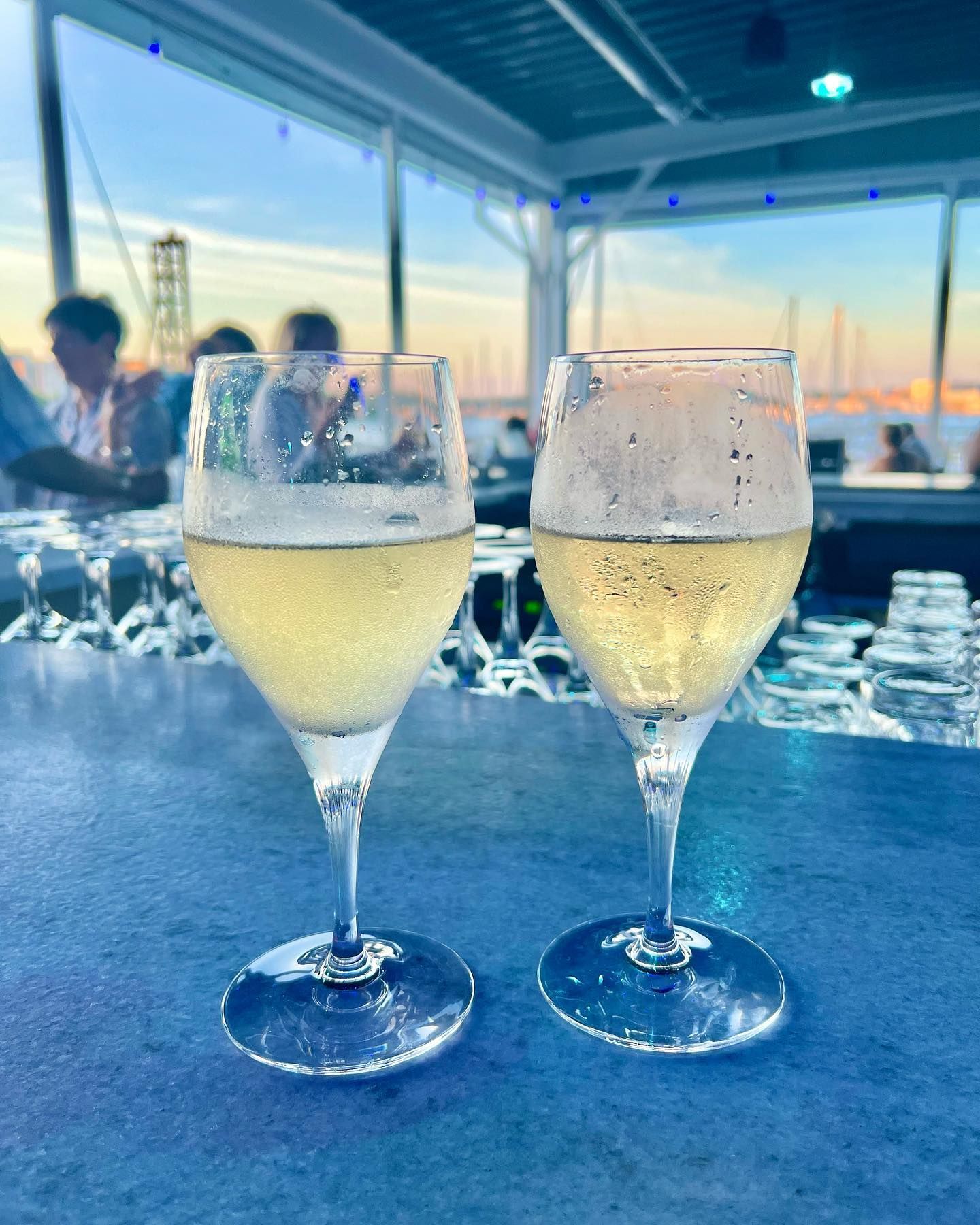Two champagne flutes filled with bubbly drink on a blue bar, waterfront view.