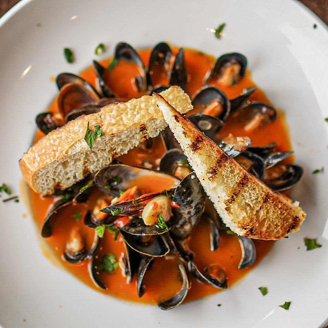 Mussels in orange sauce with toasted bread in a white bowl.