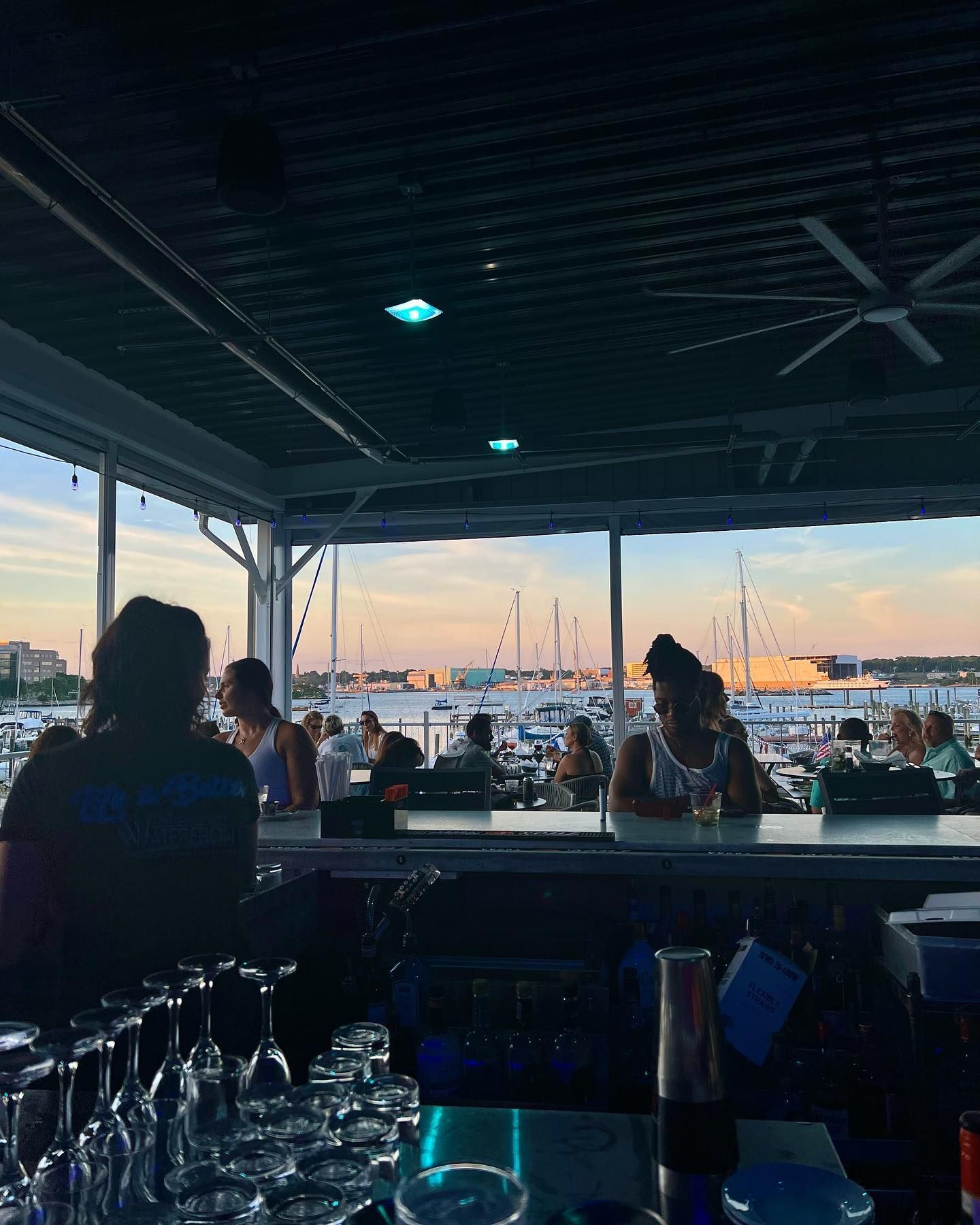 Bar patrons overlooking a marina at sunset. Silhouetted figures at bar, boats in background.