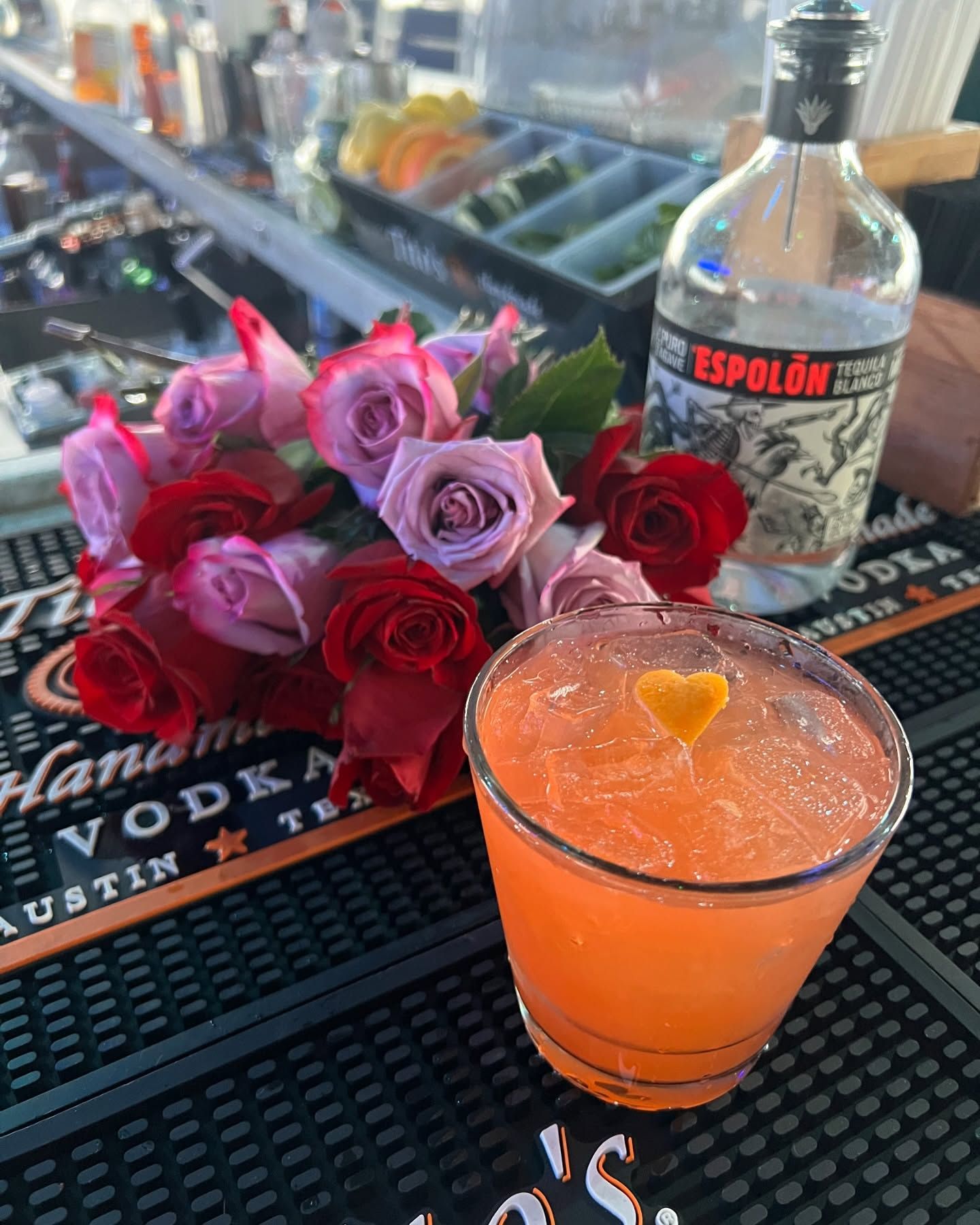 Cocktail with orange garnish, roses, and tequila bottle on a bar top.