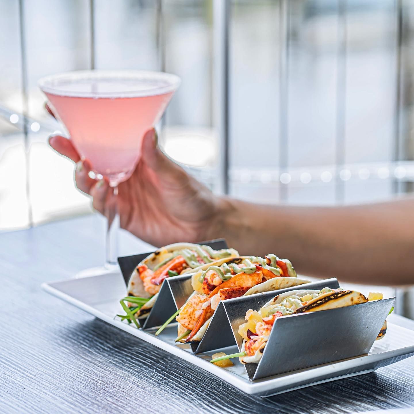 Hand holding a pink cocktail, next to a plate of lobster tacos in black holders.