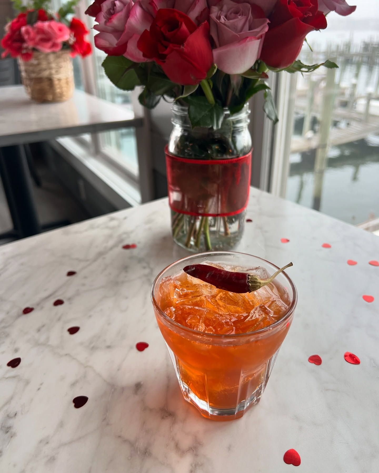 Cocktail with chili garnish on marble table with roses, heart confetti, and a window view.
