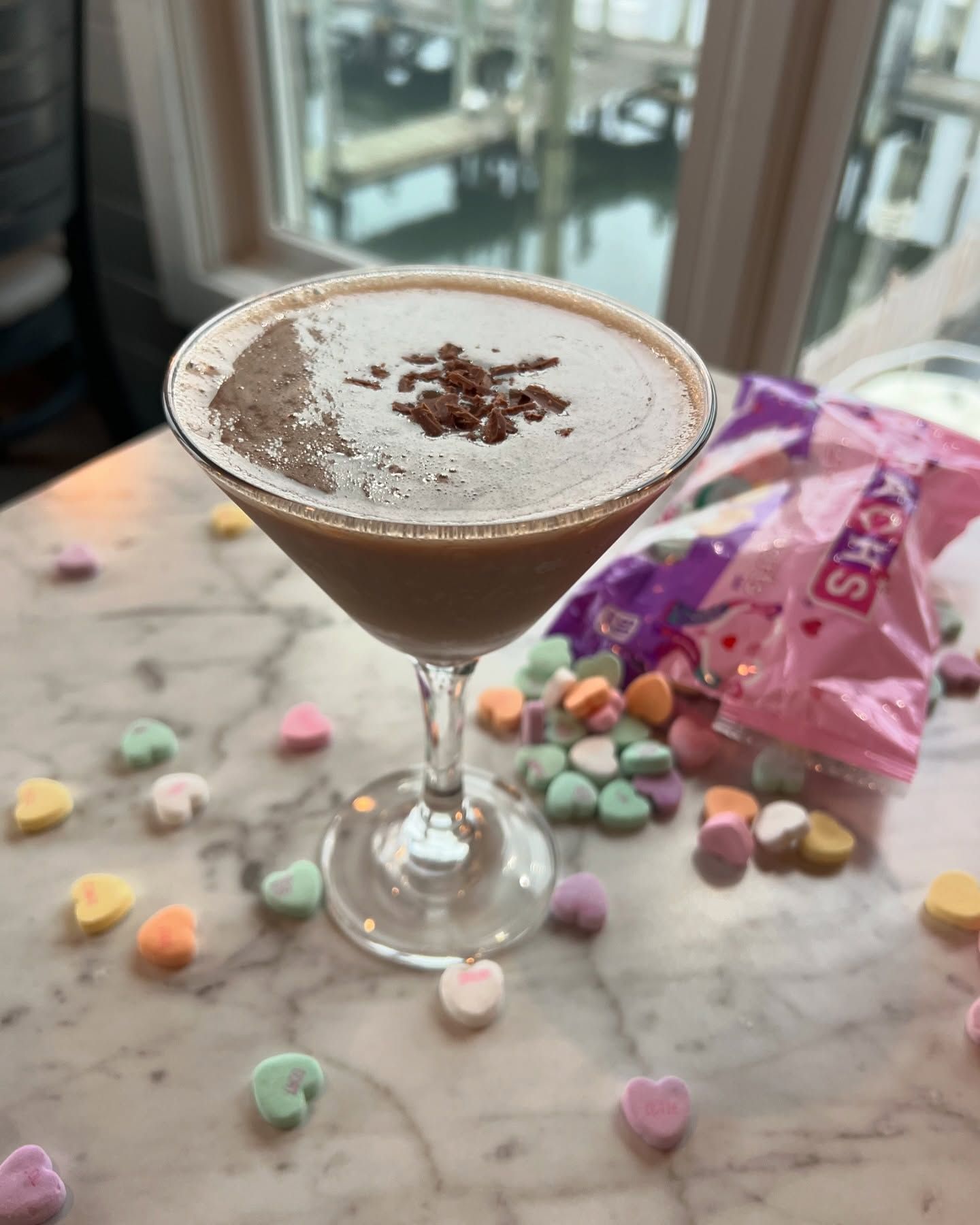 Chocolate martini, dusted with cocoa and chocolate flakes, beside scattered heart candies and a bag of conversation hearts.