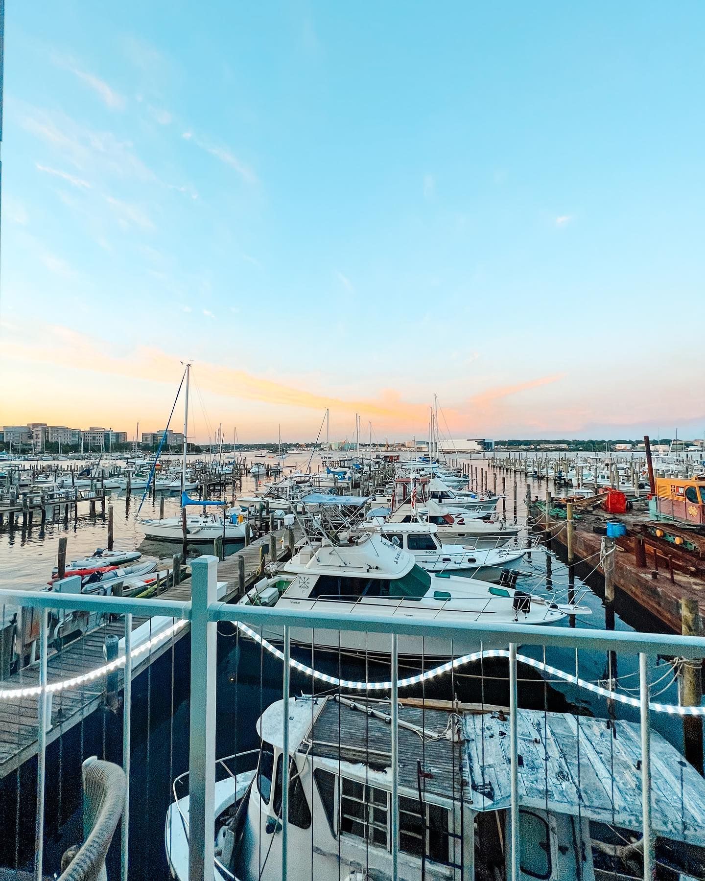 Boats docked in a marina under a colorful sunset, with a white railing in the foreground.