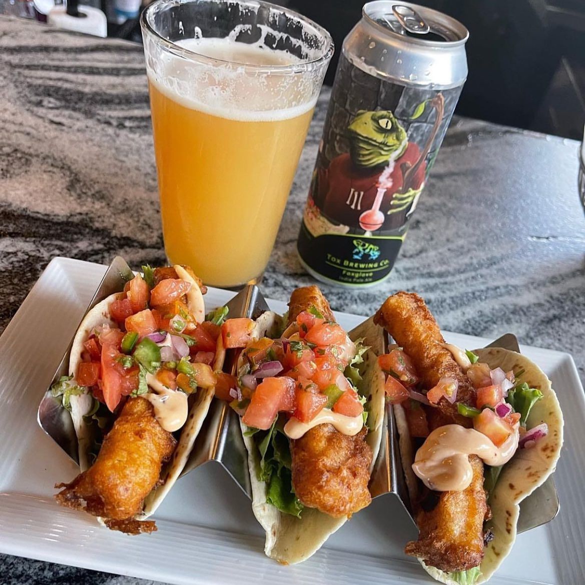 Three tacos on a plate with beer and a can. Tacos have fried food, salsa, and sauce. Beer is in a glass; a can is beside it.