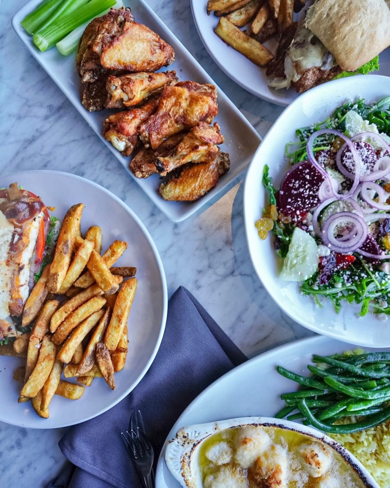 Overhead view of a table with plates of food including chicken wings, salad, sandwich, fries, and beans.