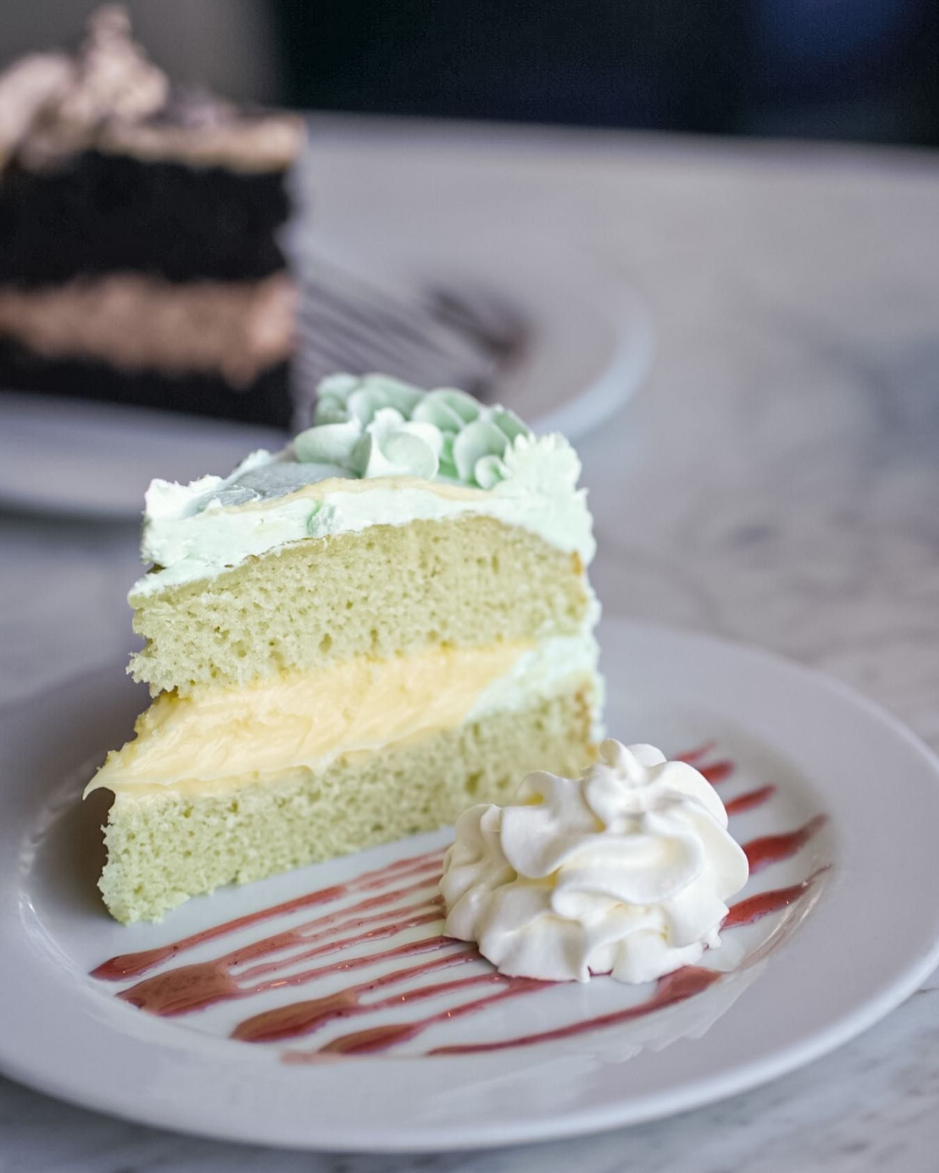 Slice of green layered cake with whipped cream on white plate. Another cake slice in the blurred background.