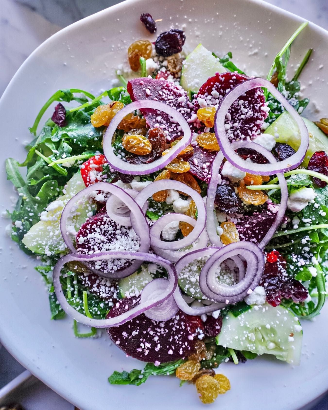 Salad with arugula, beets, red onion rings, golden raisins, goat cheese, and cucumber in white bowl.