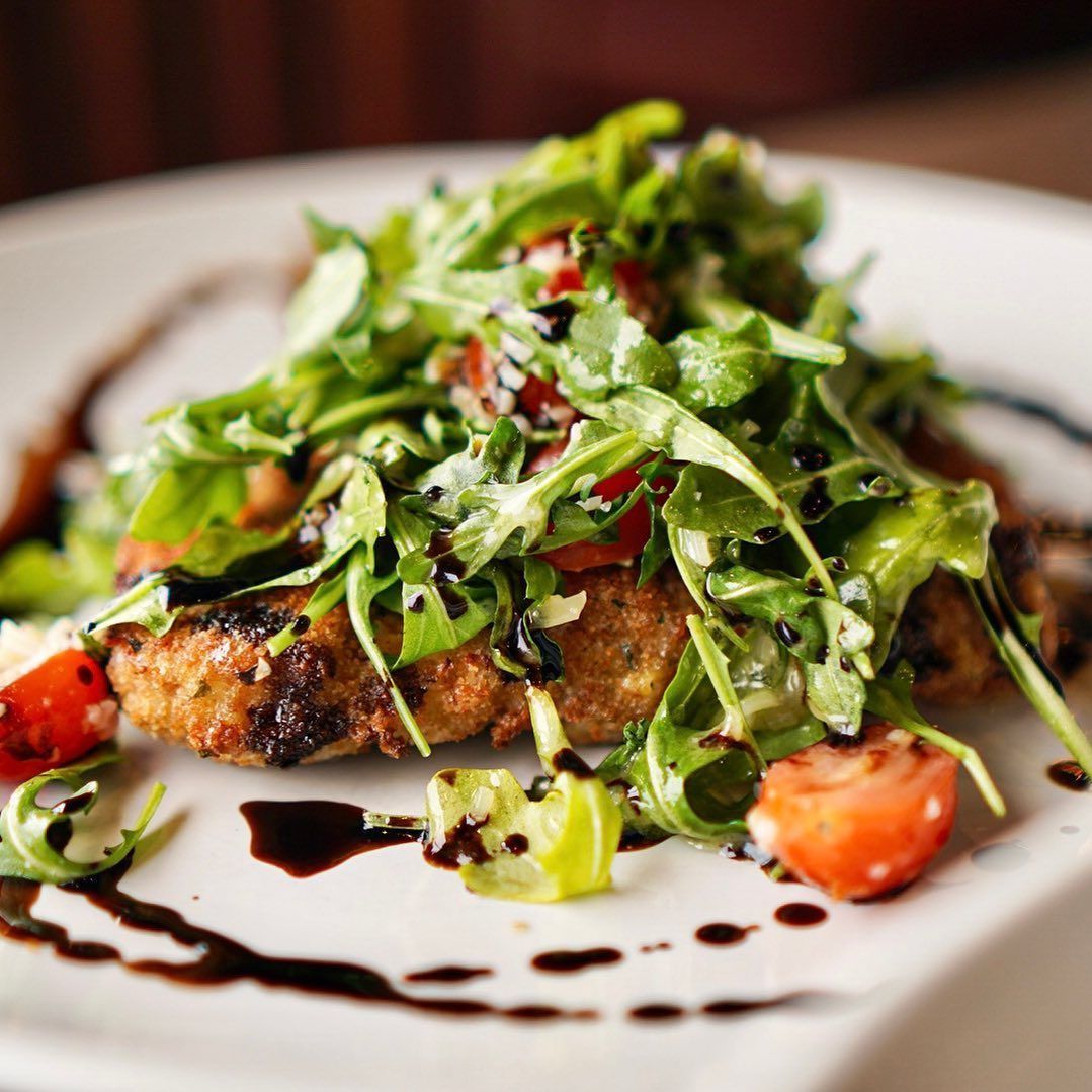 Breaded chicken cutlet topped with arugula, tomatoes, and balsamic glaze on a white plate.