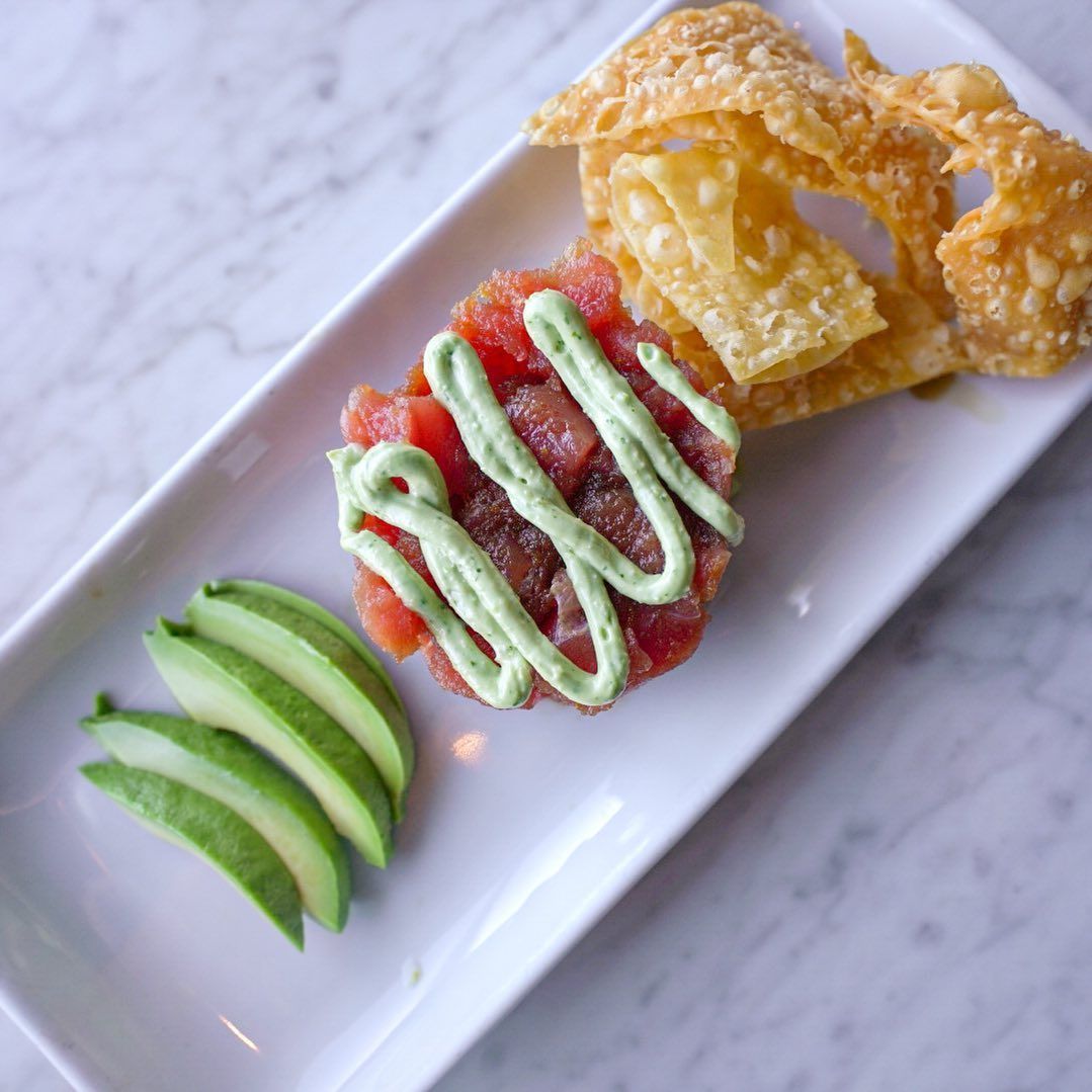 Plate of tuna tartare with avocado slices and wonton chips, drizzled with green sauce.