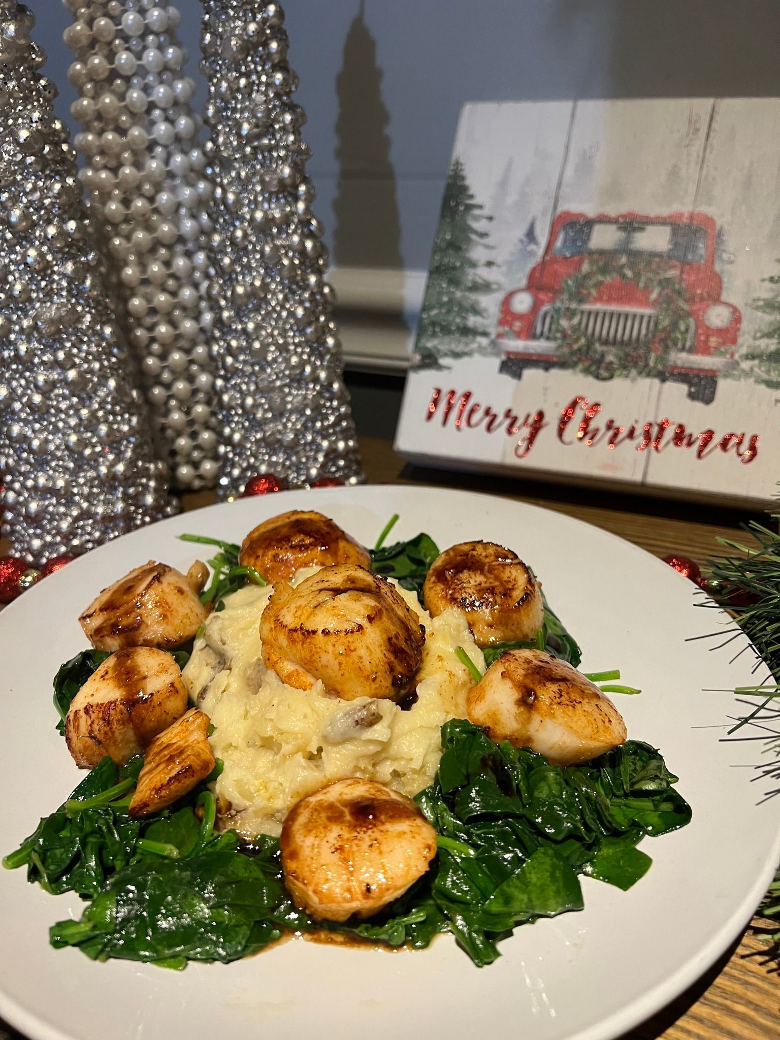 Seared scallops with spinach and mashed potatoes, garnished with glaze. Christmas decorations in the background.
