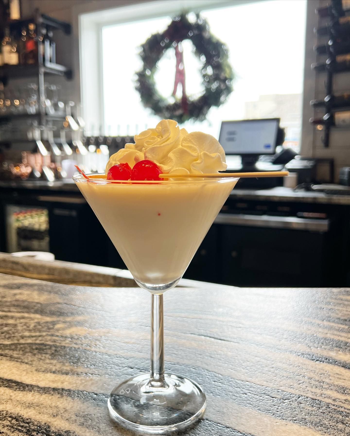 Martini glass with creamy cocktail, topped with whipped cream and a cherry, set on a bar.