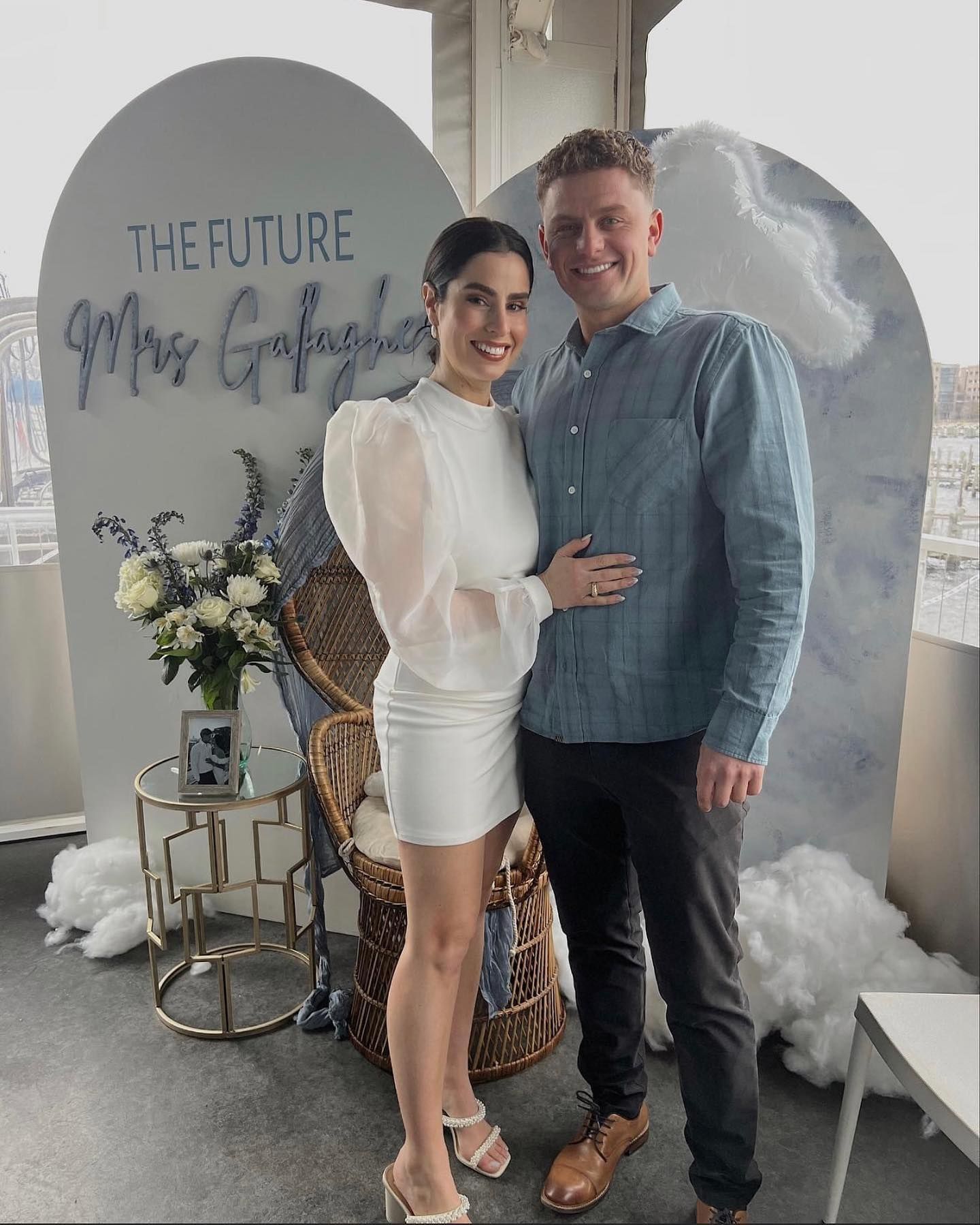 Couple posing near a backdrop with clouds and text. Woman in white dress, man in blue shirt, smiling.