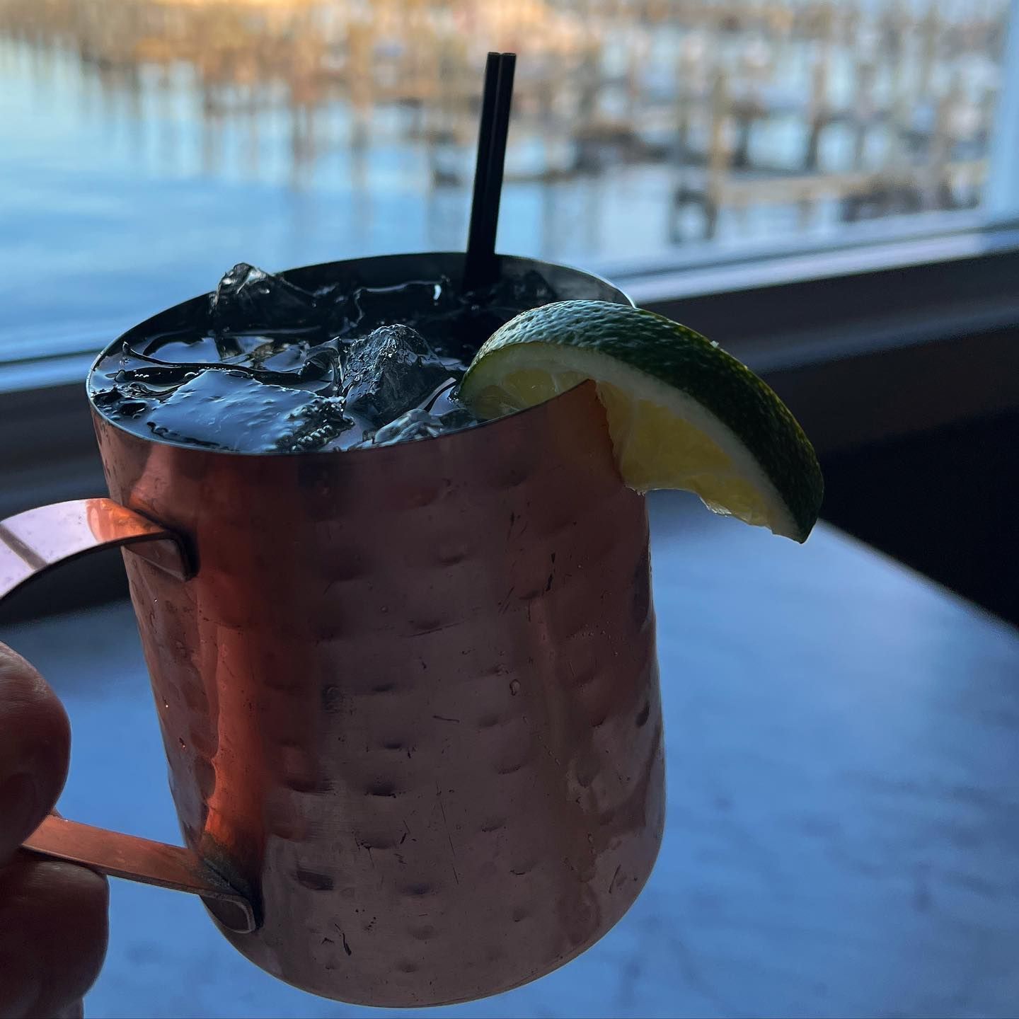 Copper mug with a Moscow Mule cocktail, lime garnish, and black straw. Waterfront view.