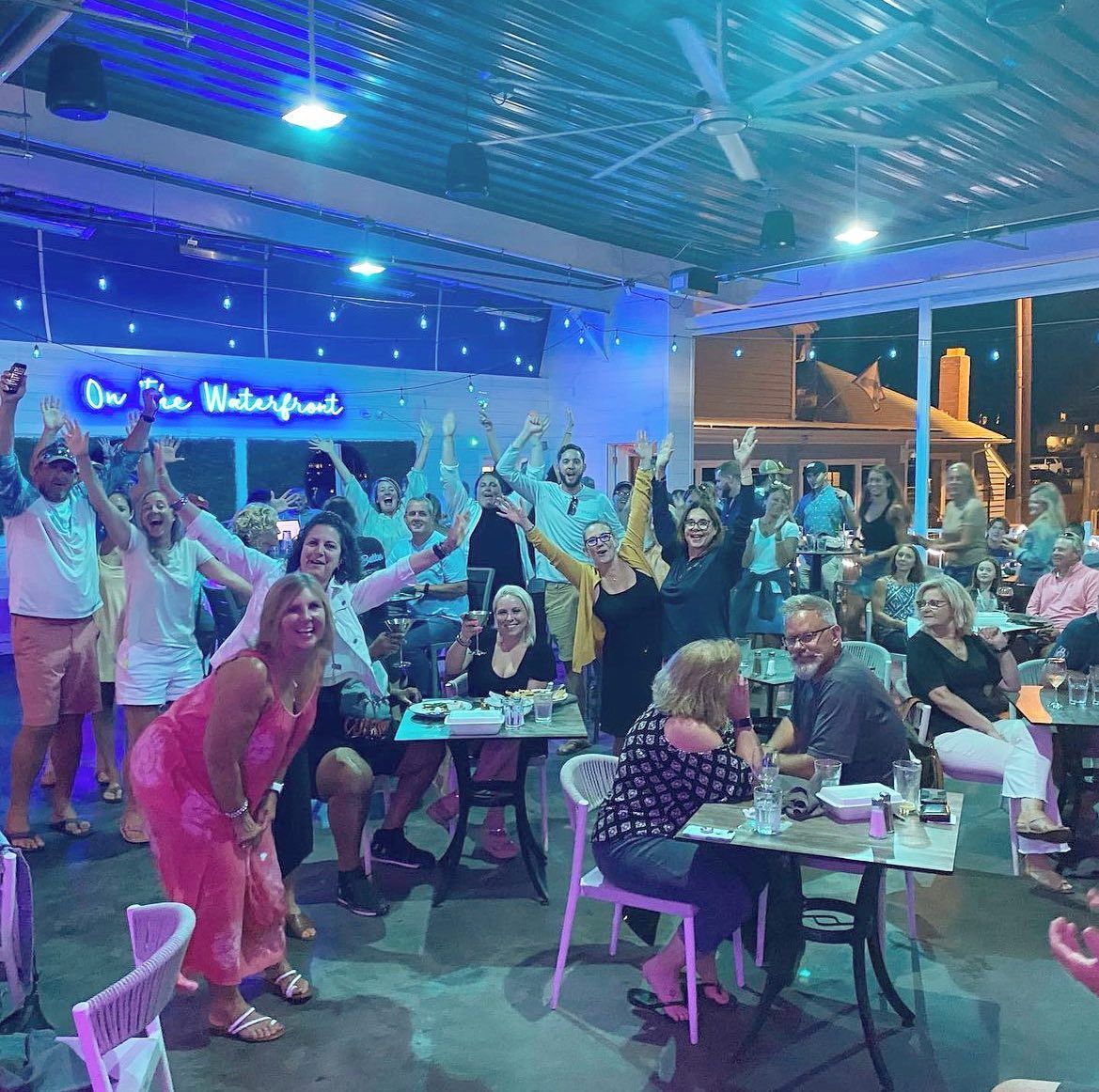 People at a waterfront bar celebrating with arms raised. Blue and white decor, tables and chairs visible.