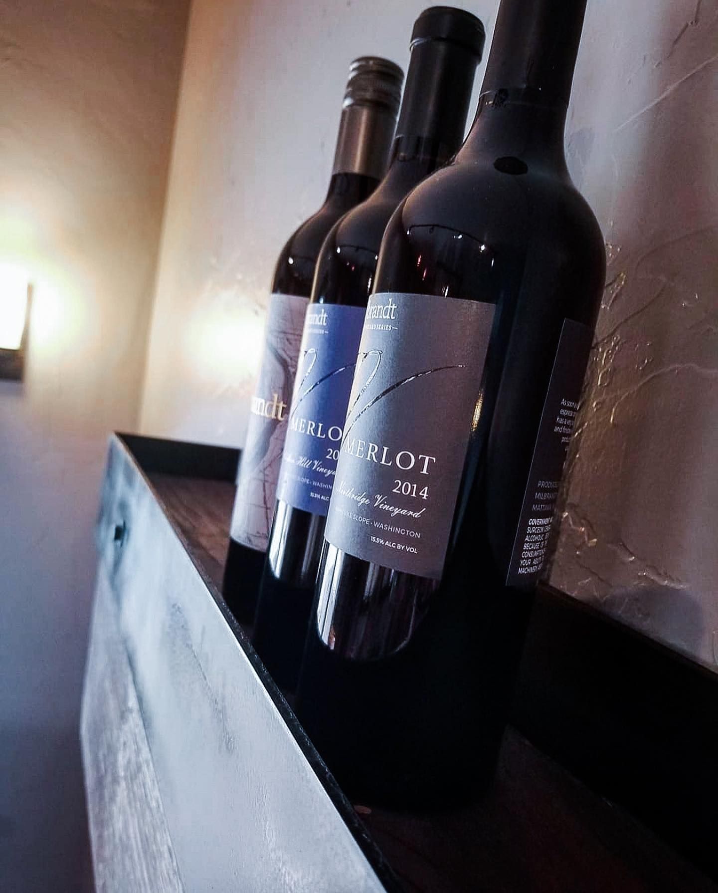 Three wine bottles on a shelf, lit from the side. The middle bottle is labeled Merlot.