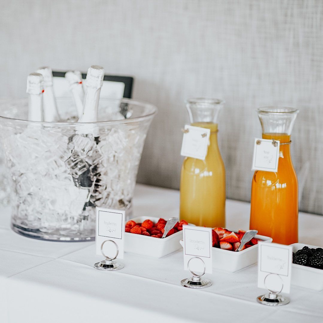 Champagne bottles in ice bucket, juice, berries, and signs on white table.