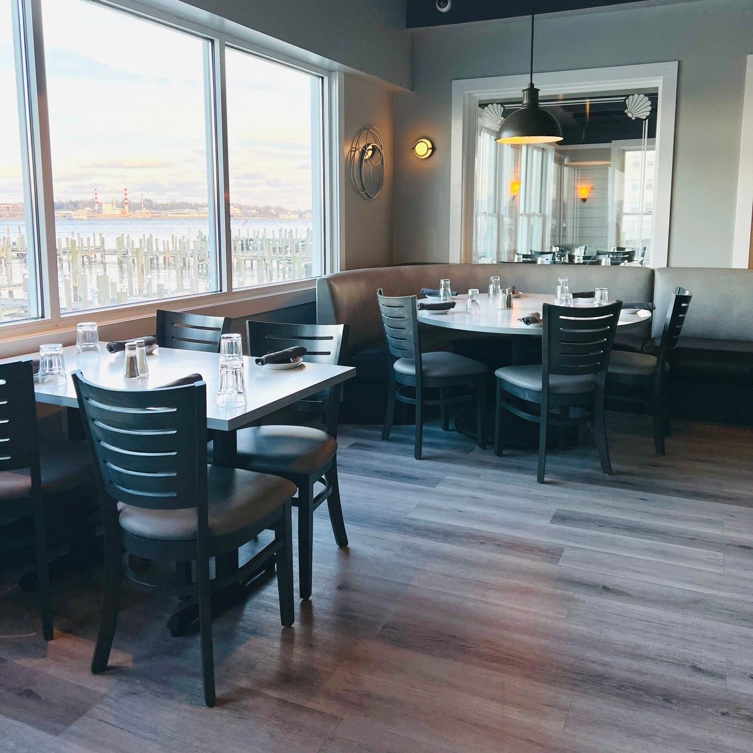 Restaurant interior with tables and chairs, large windows with a harbor view, and neutral decor.