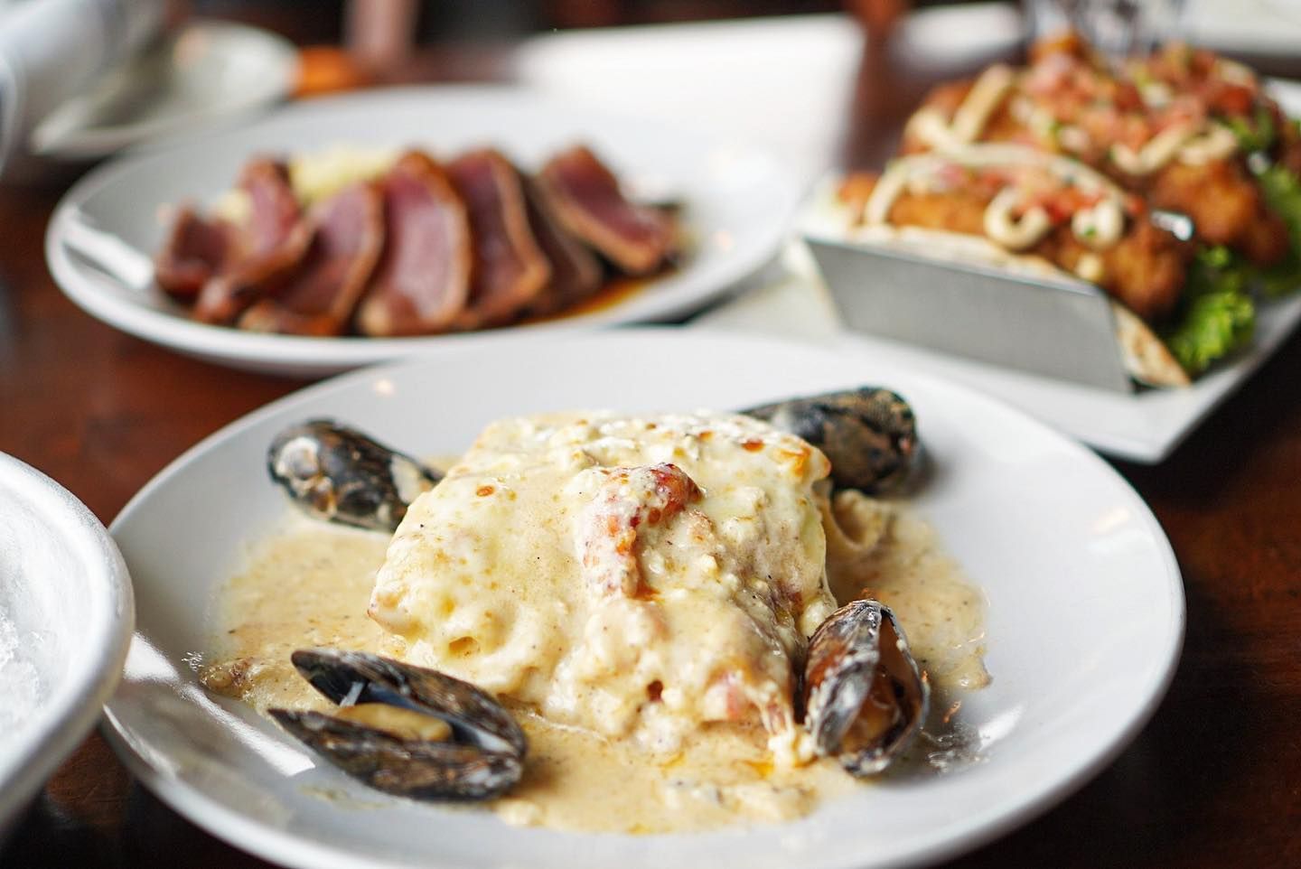 Seafood dishes on white plates seared tuna, pasta with mussels, and tacos on a dark wooden table.