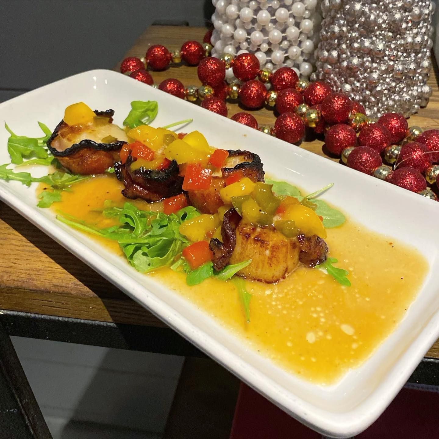 Seared scallops on arugula with mango salsa and sauce, presented on a white plate. Festive red and gold beads in the background.