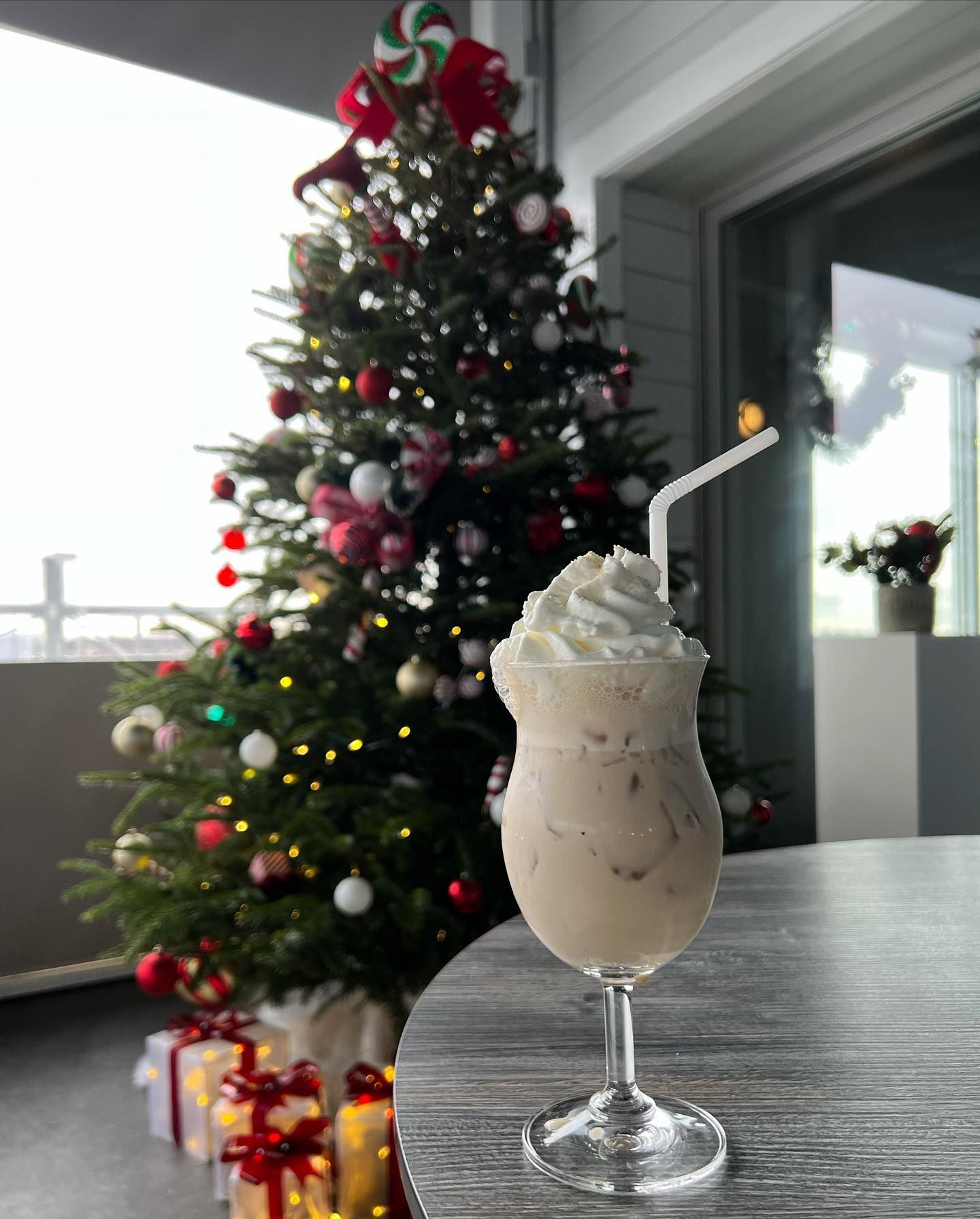 A festive milkshake with whipped cream and straw sits on a table near a decorated Christmas tree with gifts.