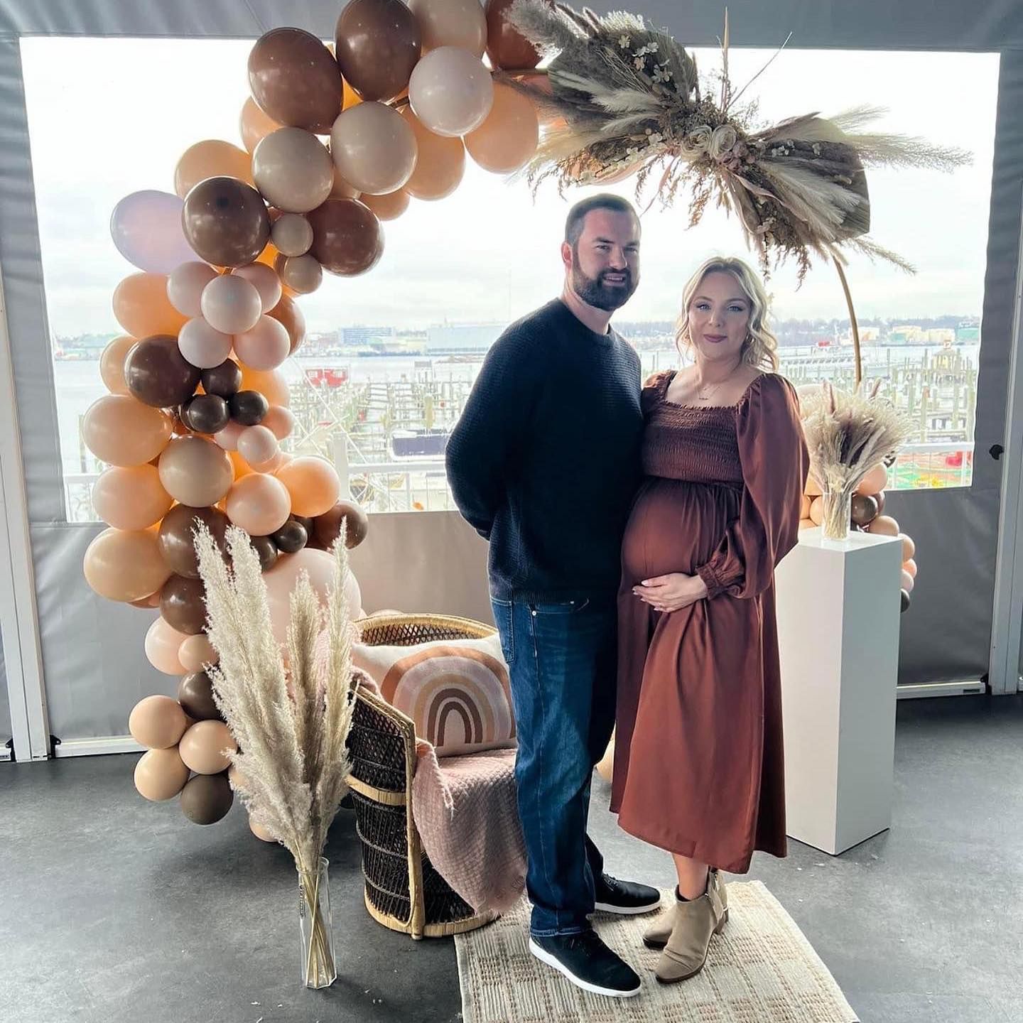 Couple at a baby shower with neutral balloons, bohemian decor, and city view. Woman cradles her belly.