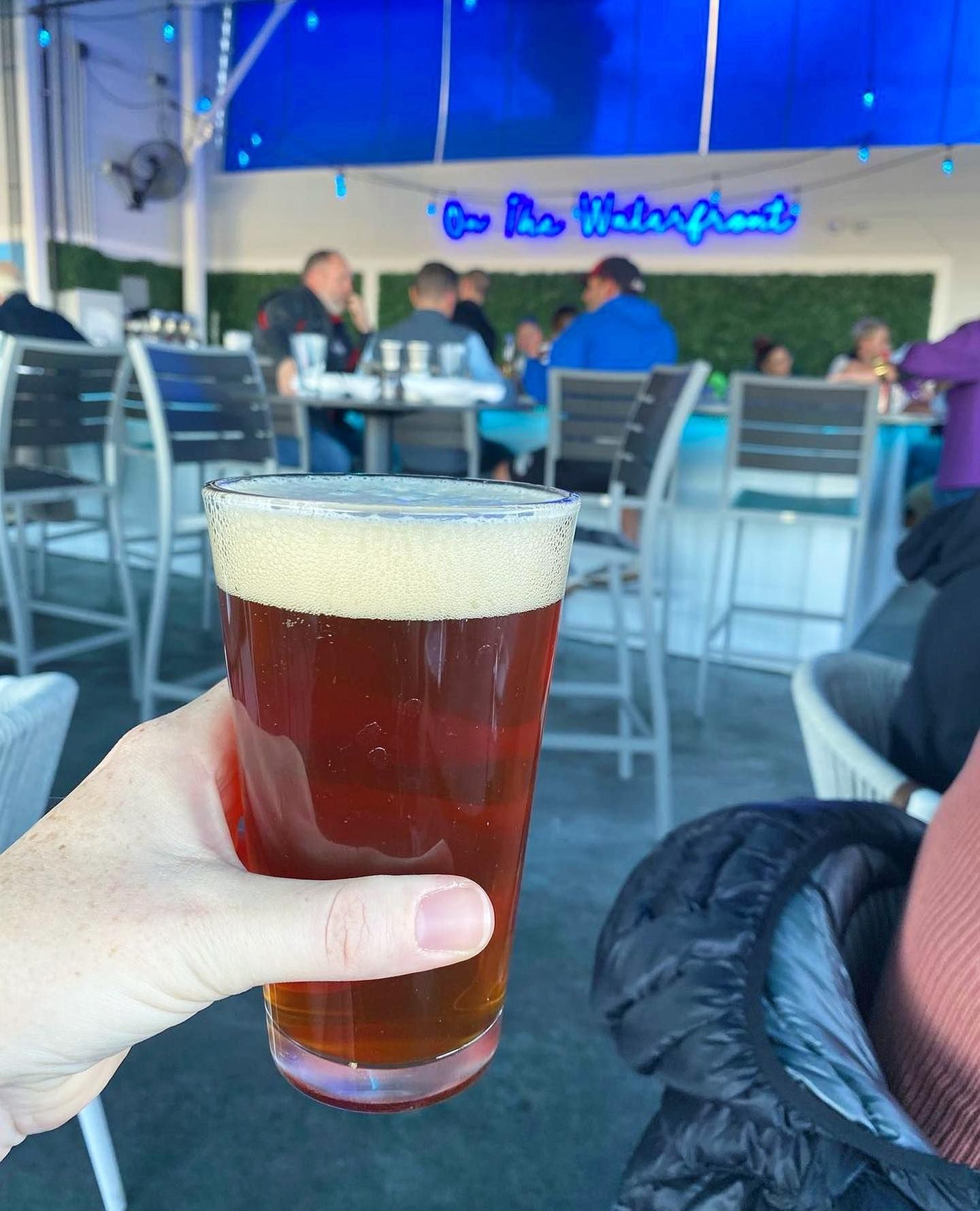 Hand holding a pint of beer at an outdoor bar with patrons and a blue illuminated sign.