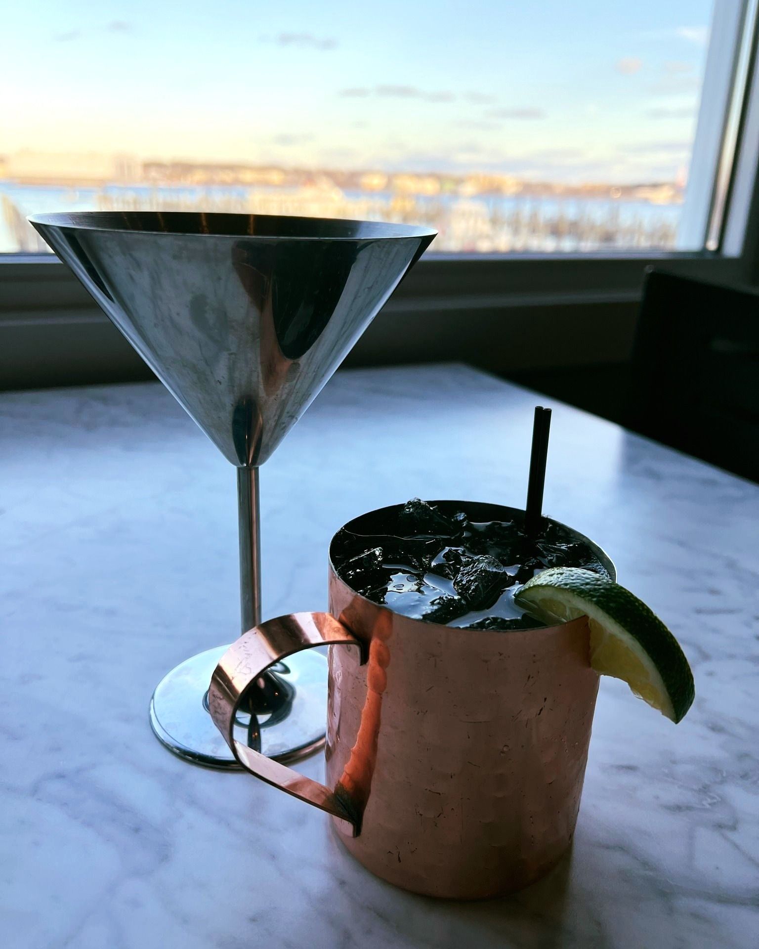 Moscow Mule in copper mug, martini glass on marble table near window with city view.