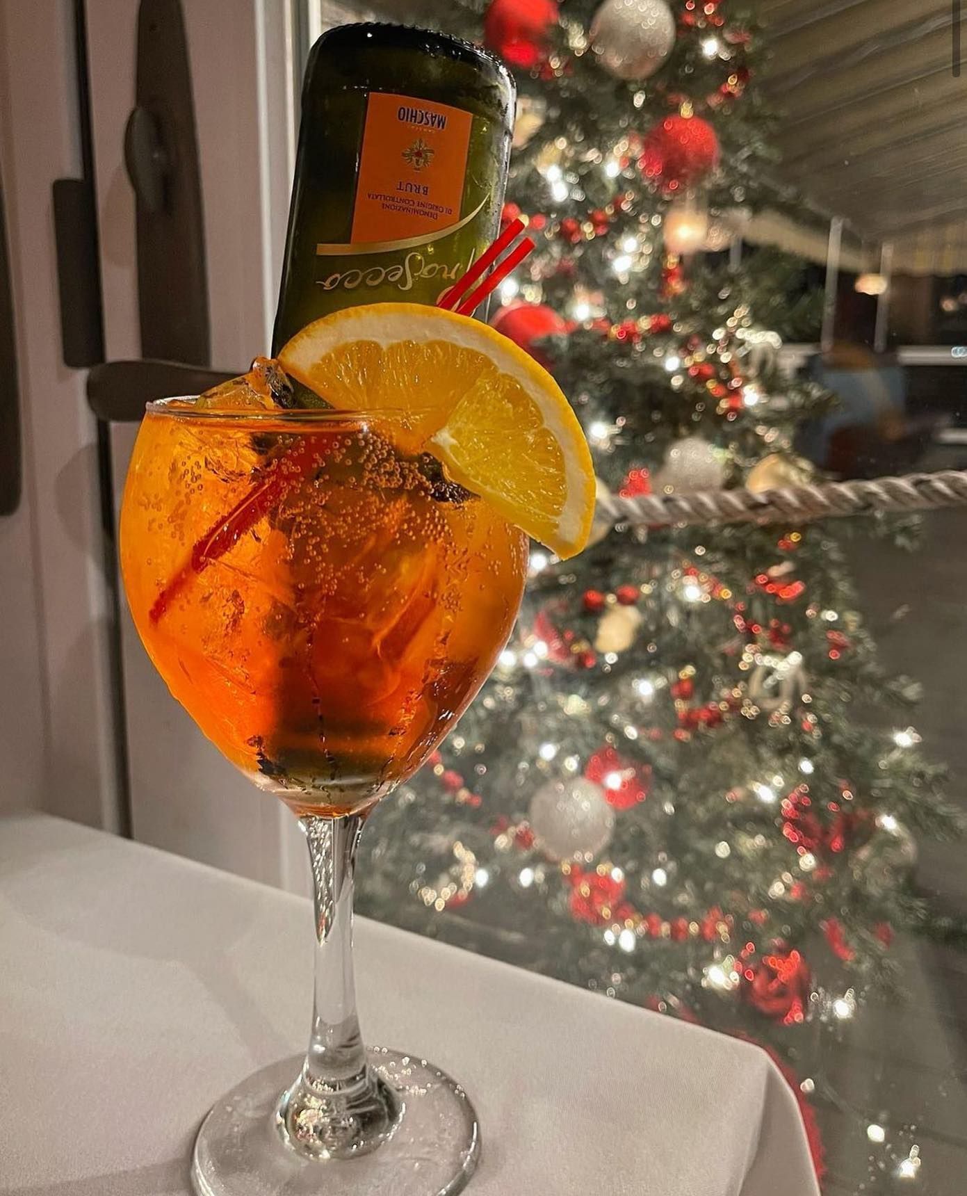 Aperol spritz with orange slice and straw, champagne bottle in a glass, Christmas tree in the background.