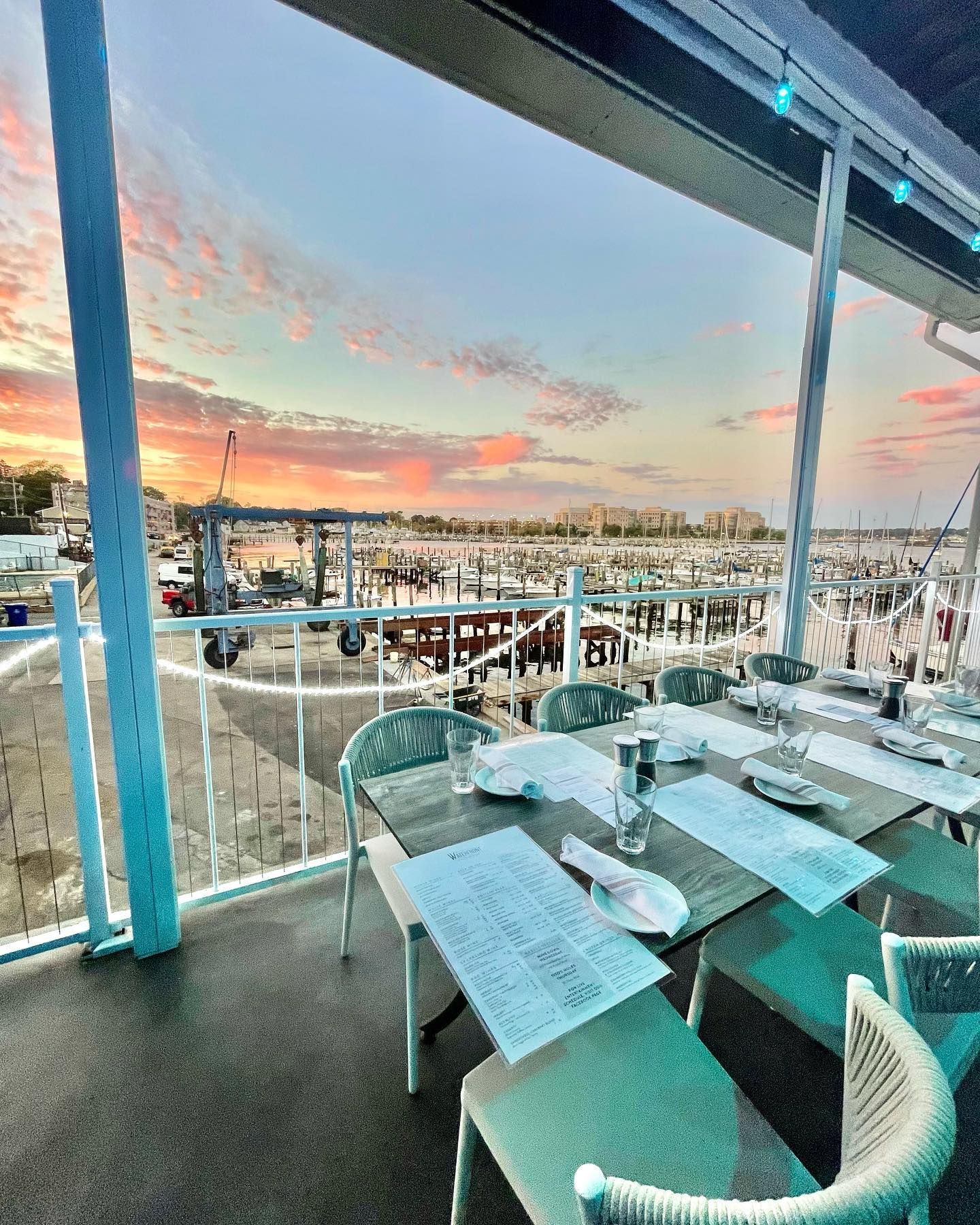Outdoor dining area overlooking a harbor at sunset. Tables set with menus, boats docked, and a colorful sky.