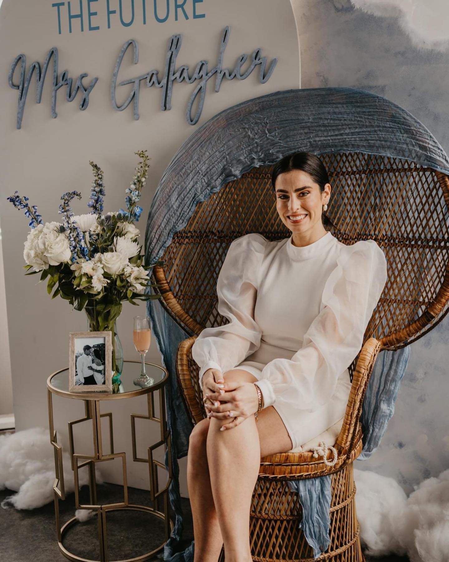 Woman in white dress sits in a peacock chair at a bridal shower, blue and white decor.