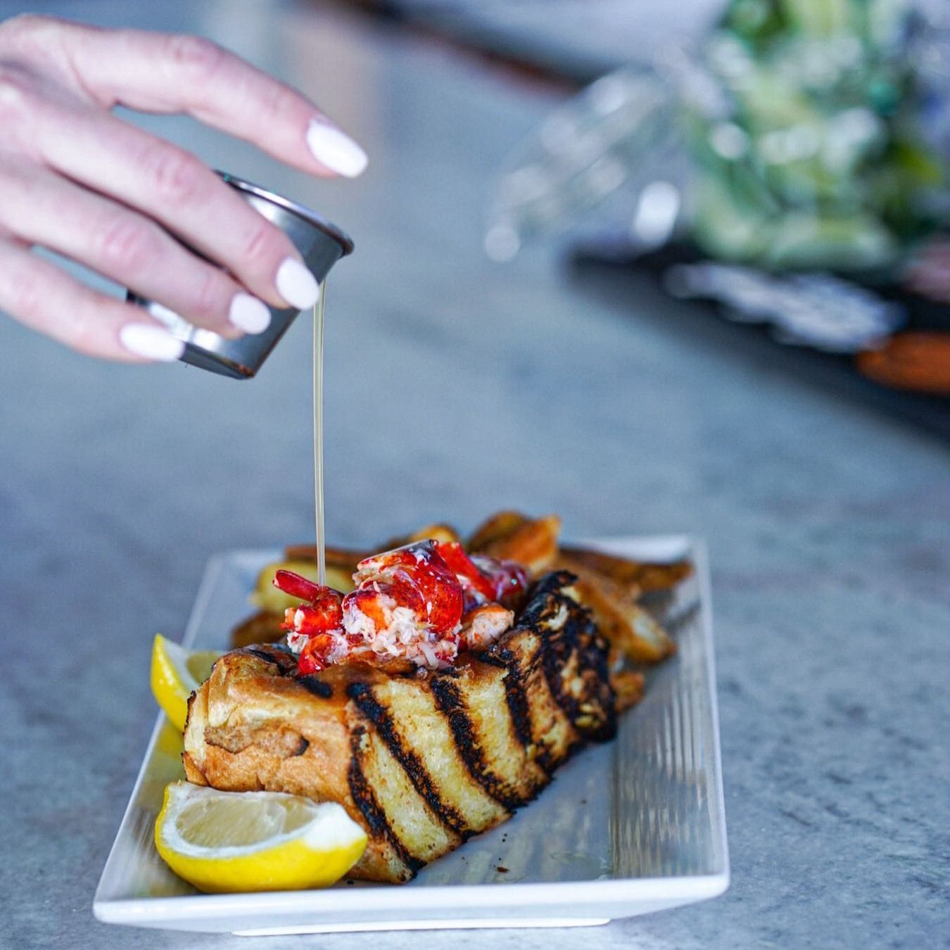 Hand pouring sauce over lobster on grilled bread with lemon slices.