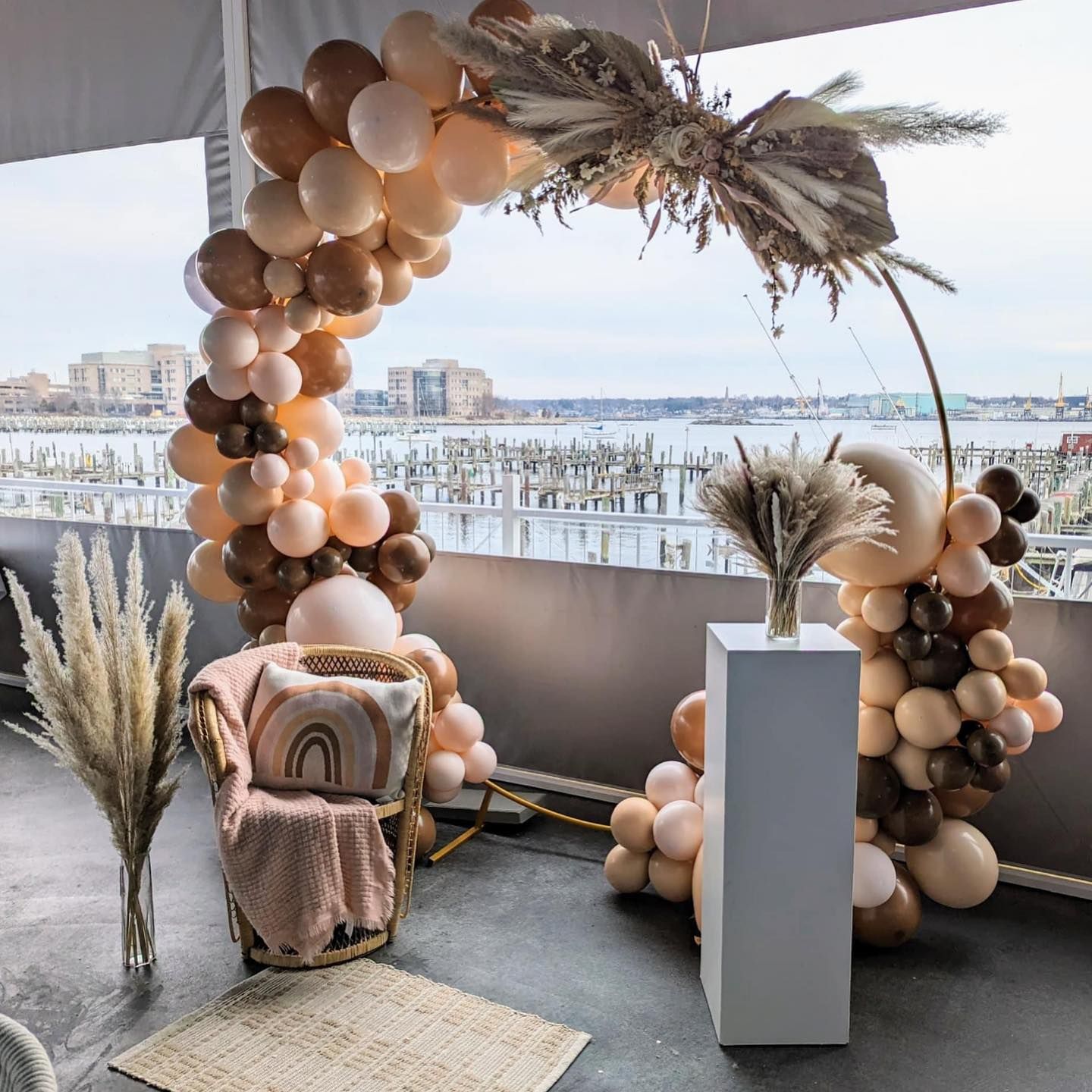 Boho-style party setup with balloon arch, chair, pampas grass, and a city waterfront backdrop.