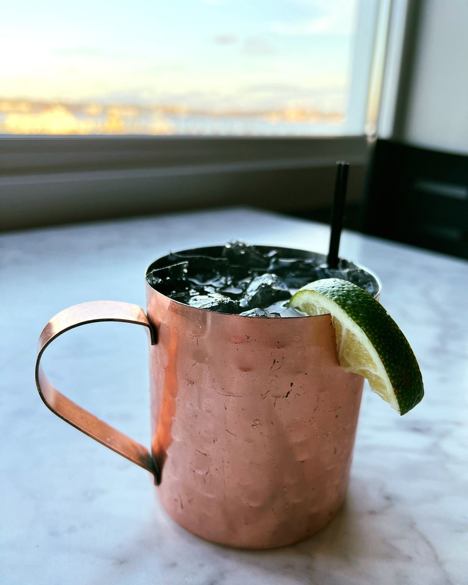 Copper mug Moscow Mule cocktail with lime and straw, on a marble table with a city view.