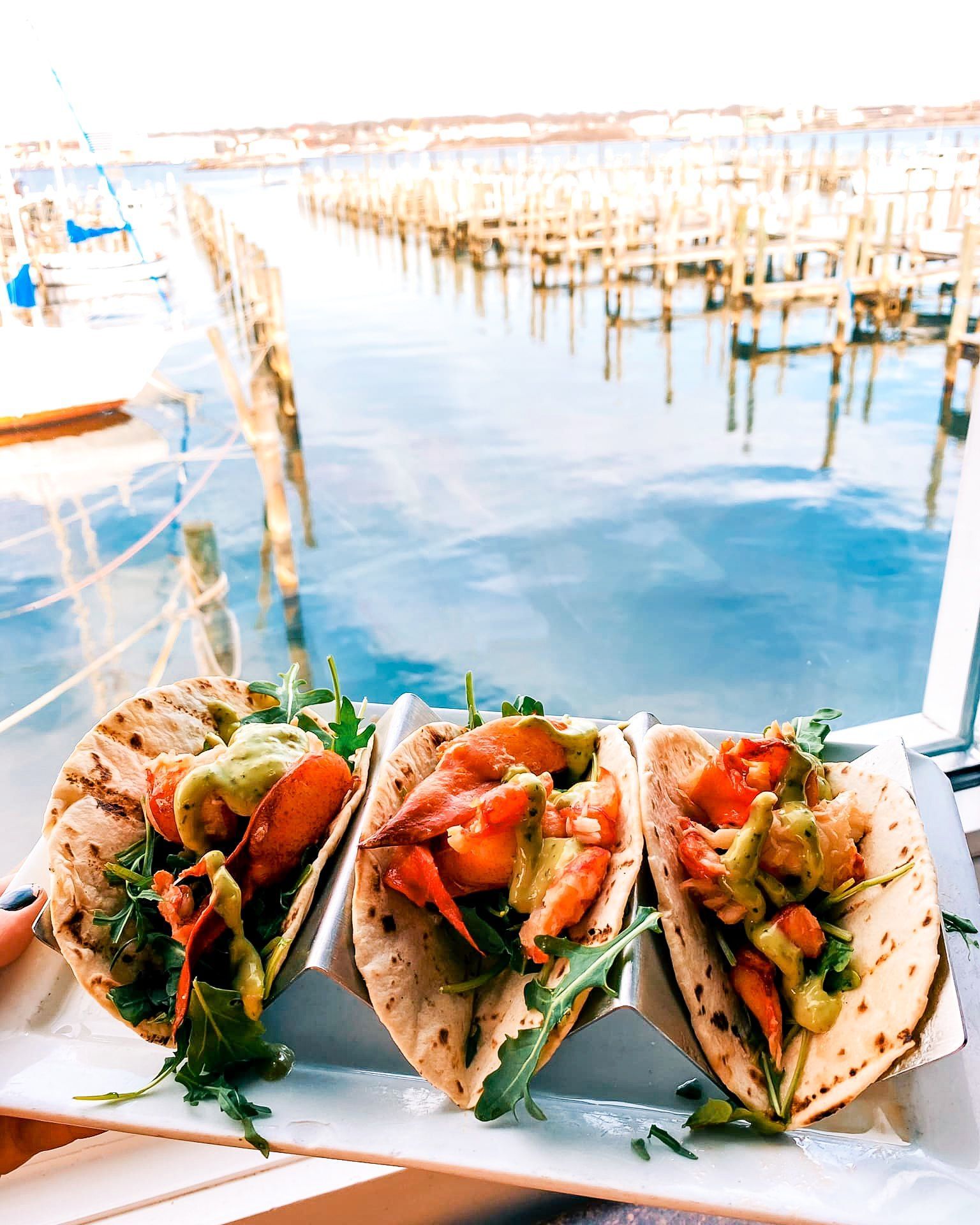 Three shrimp tacos with avocado sauce on a white plate overlooking a harbor.