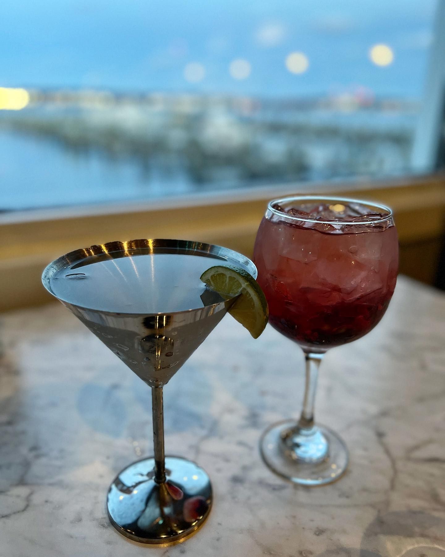Two cocktails in metallic glasses sit on a marble surface near a window with an outdoor view.