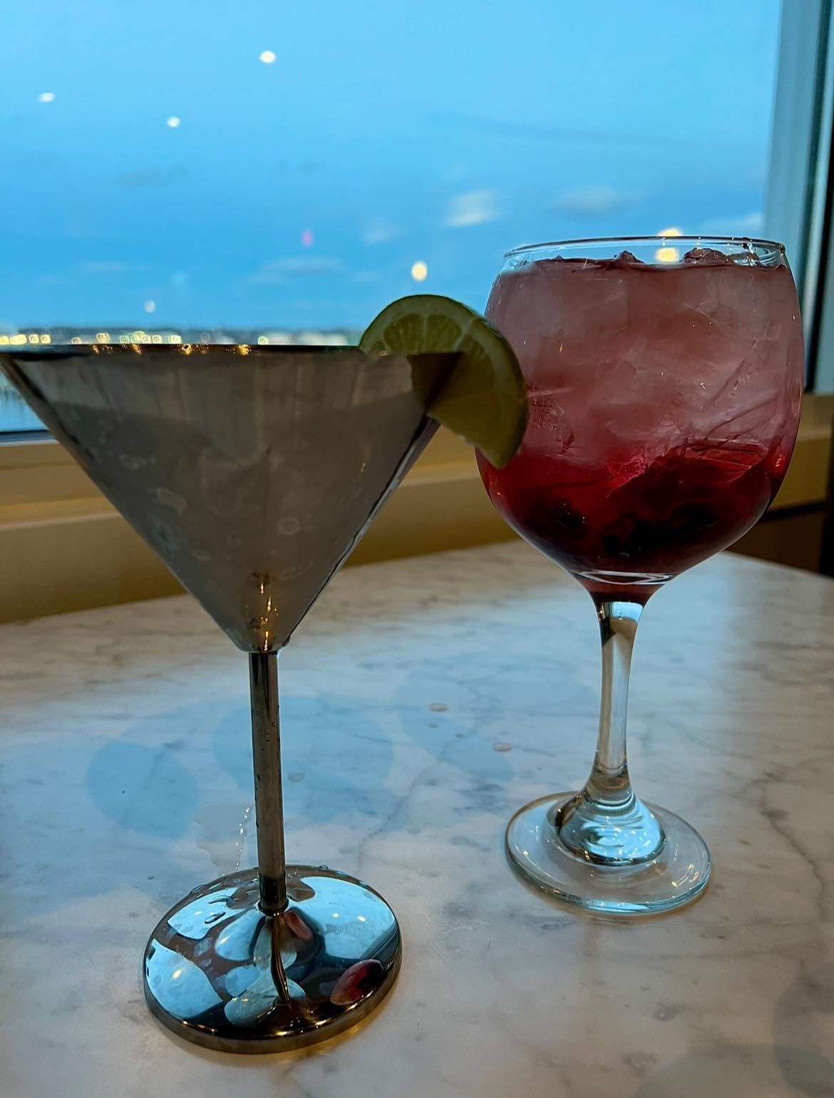 Two cocktails in silver glasses, one with lime, on a table near a window.