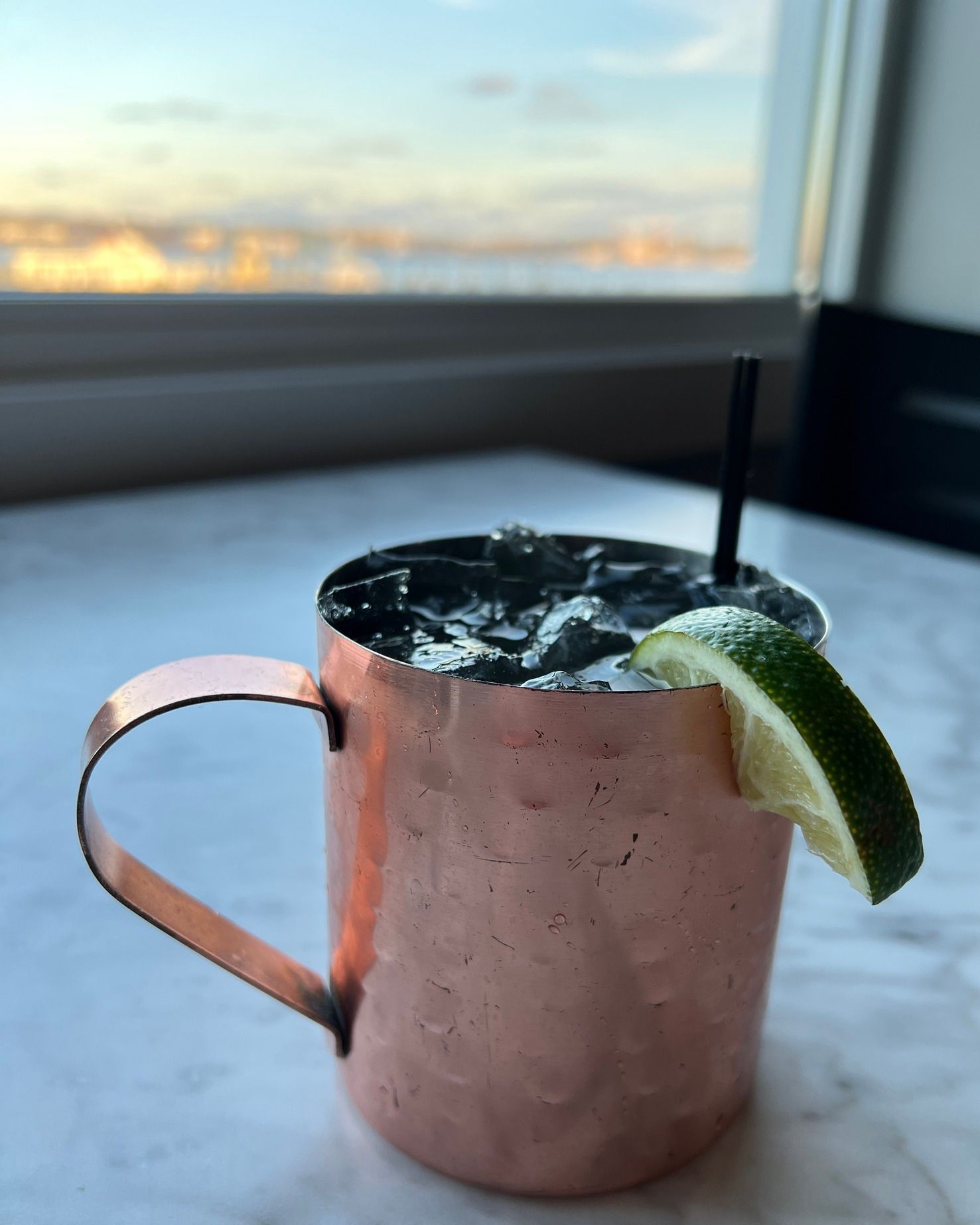 Copper mug cocktail with lime, ice, and straw on marble, window in background.