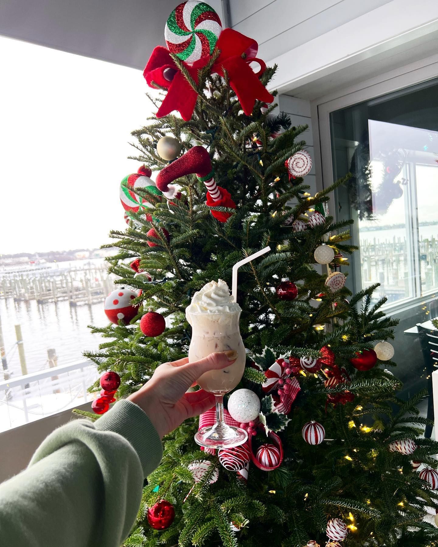 Hand holding a drink in front of a decorated Christmas tree with outdoor view.