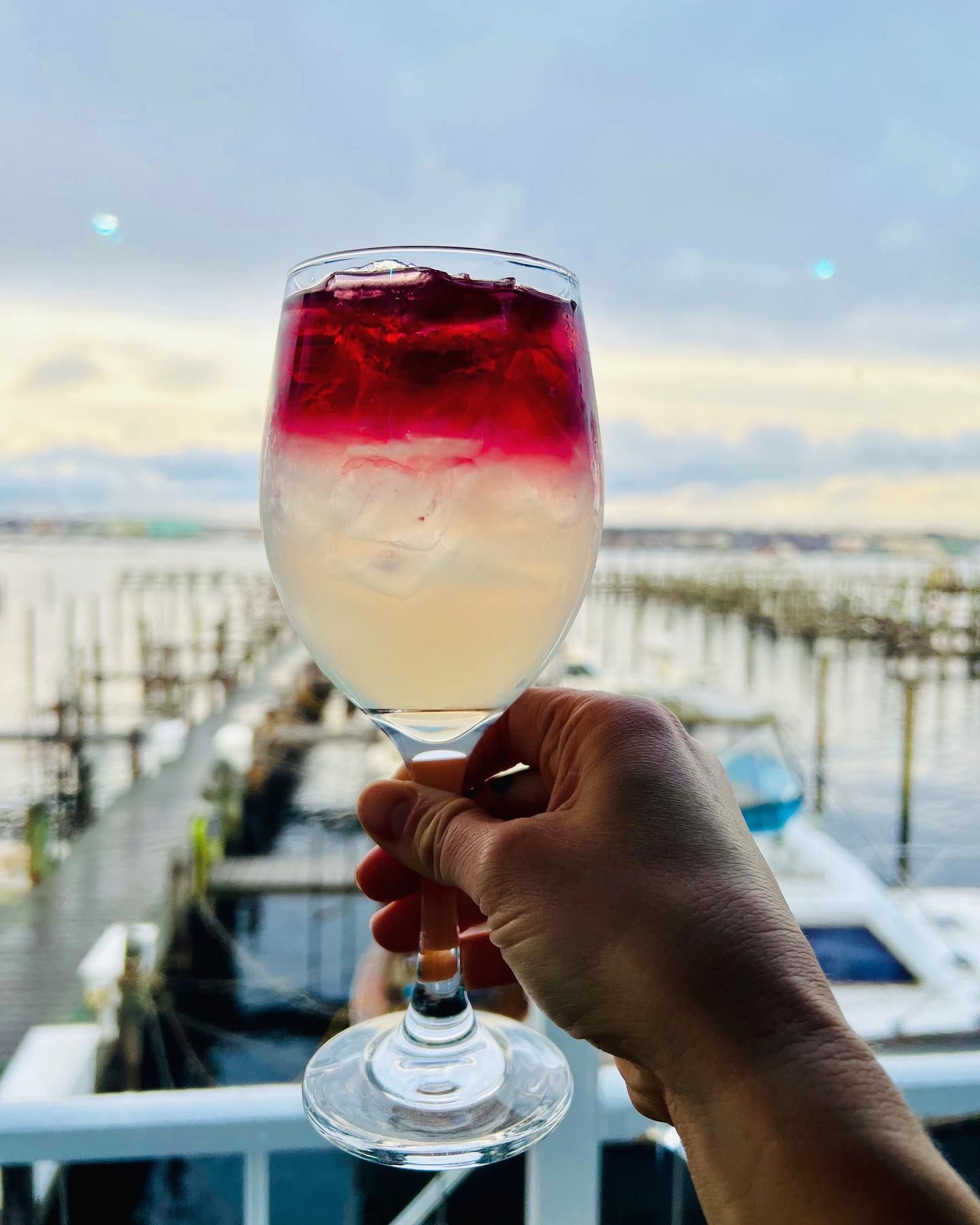 Hand holding a layered cocktail, reddish-purple over a light yellow, with a waterfront background.