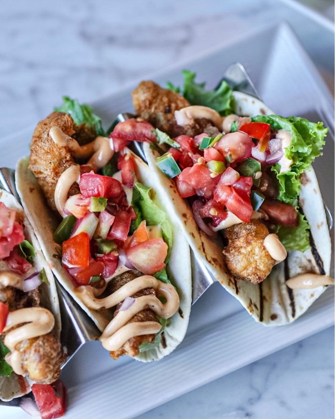 Three tacos in metal holders on a white plate. Each taco has fried food, lettuce, salsa, avocado, and sauce.