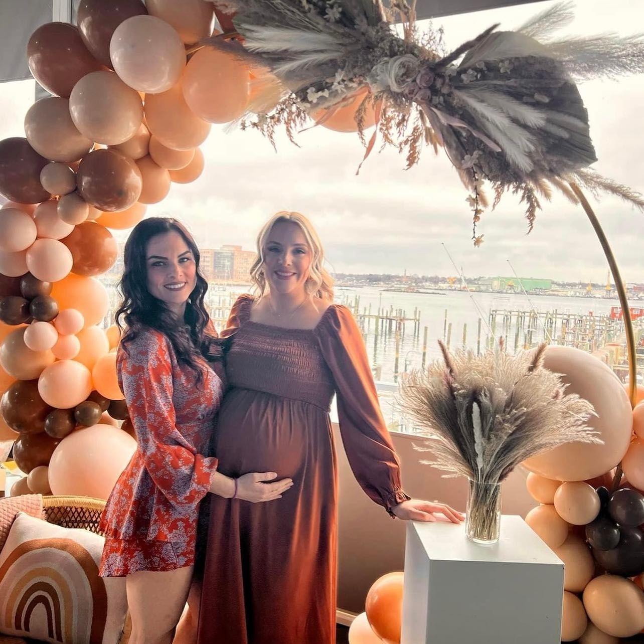 Two women at a baby shower. One pregnant, they pose near balloons, overlooking a body of water.