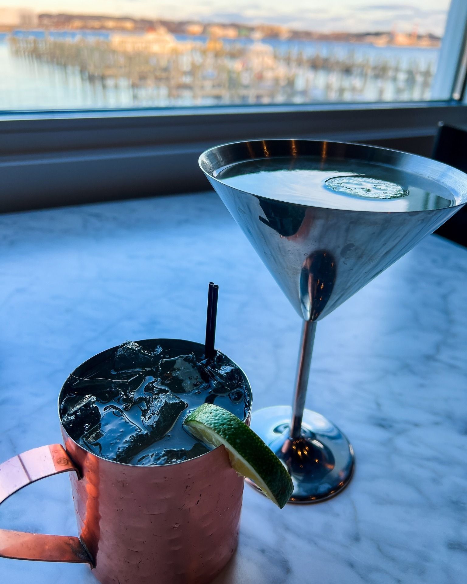 Copper mug with cocktail and lime wedge, martini glass, waterfront view.
