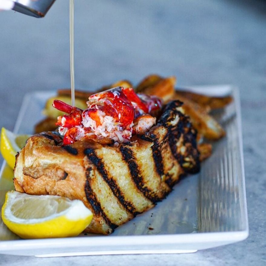 Grilled lobster roll with lobster meat, lemon, and fries, drizzled with sauce on a plate.