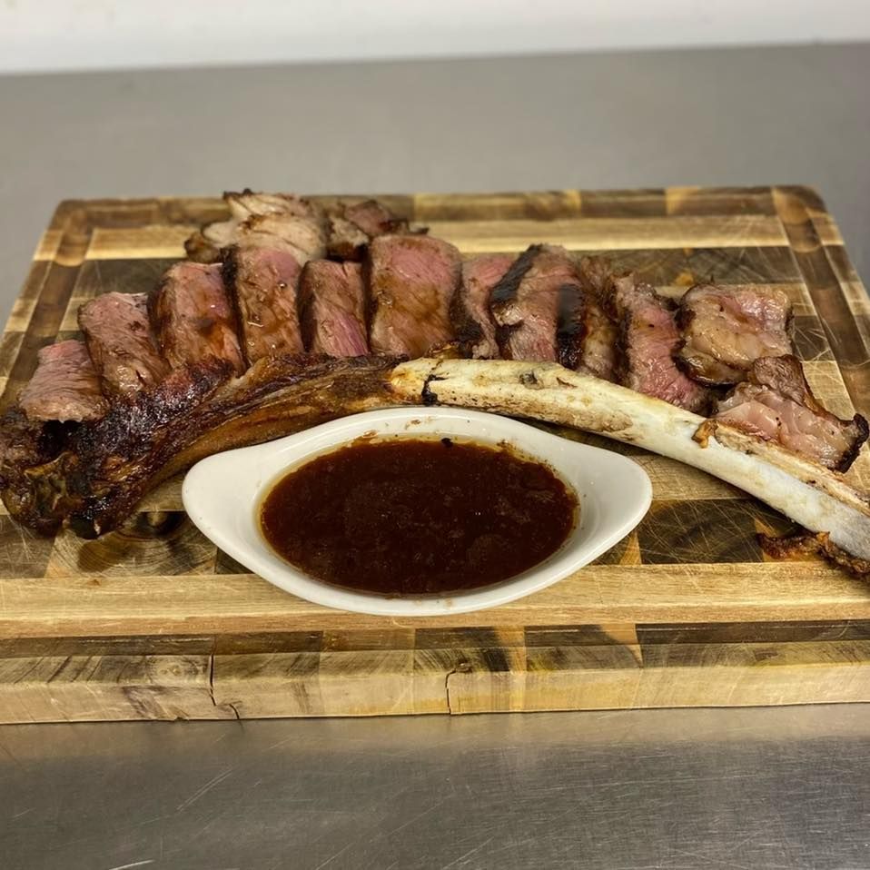 Sliced steak with bone, presented on a wooden board with dipping sauce.